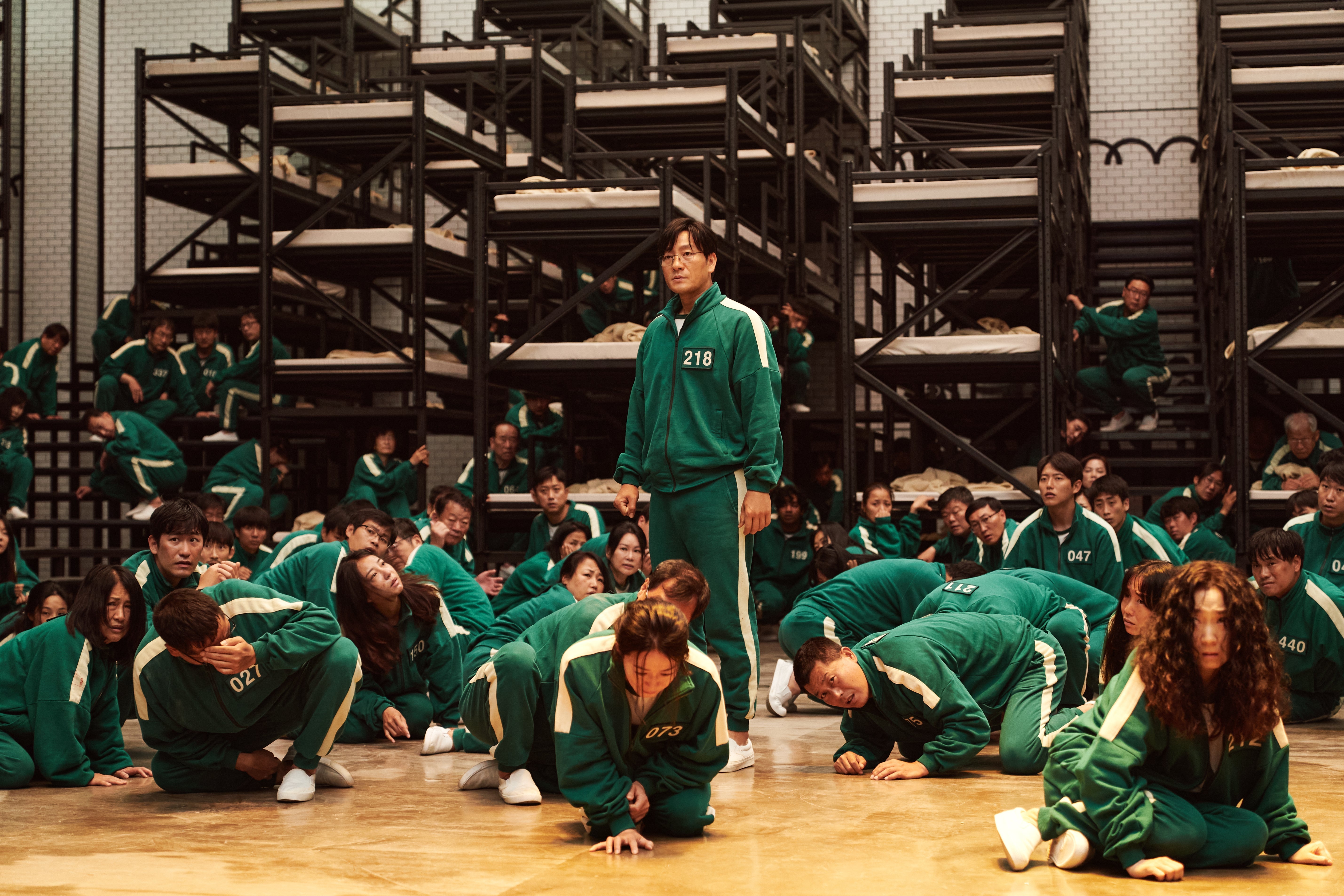 In a bunk bed-filled room, people wearing green and white jumpsuits crouch and sit on the floor. One man with the number 218 on his uniform stands upright.