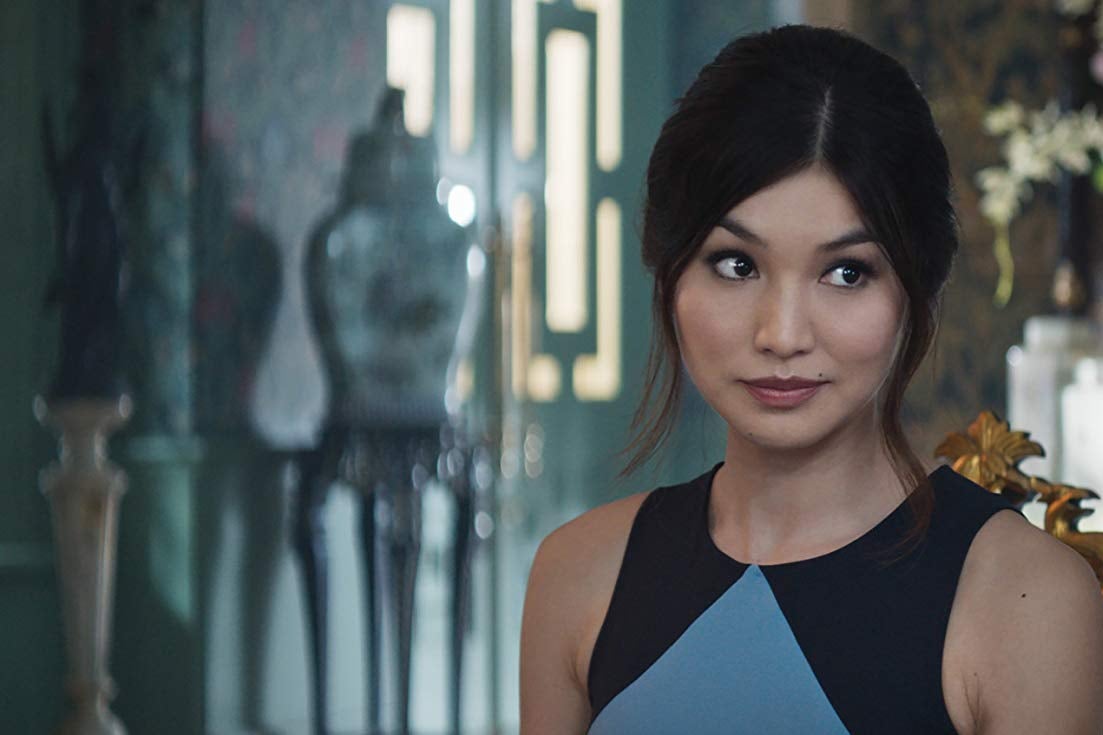 Gemma Chan wears her hair up with a few tendrils escaping. She sits with a vase and flowers on an opulent table behind her.