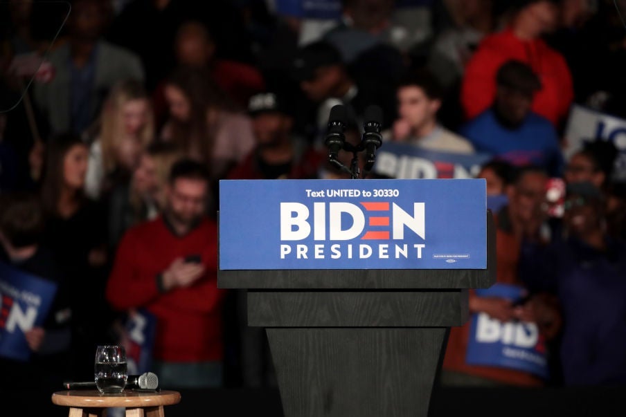 An empty lectern decorated with a Biden logo against the backdrop of supporters at a rally.