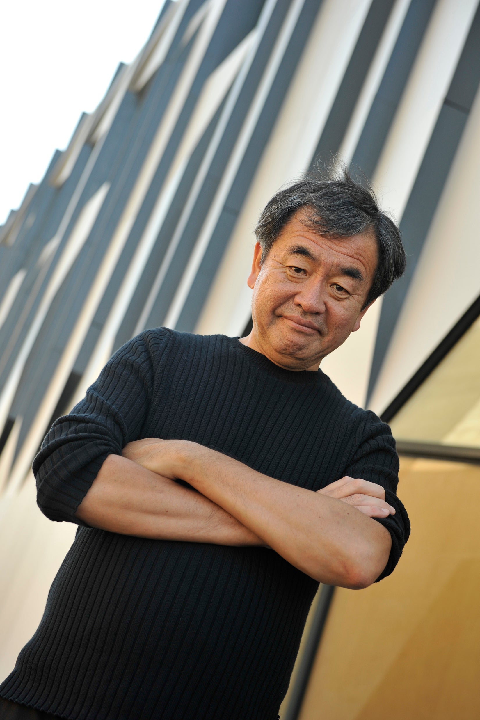 A portrait of Kengo Kuma, standing with his arms crossed and wearing a black shirt.