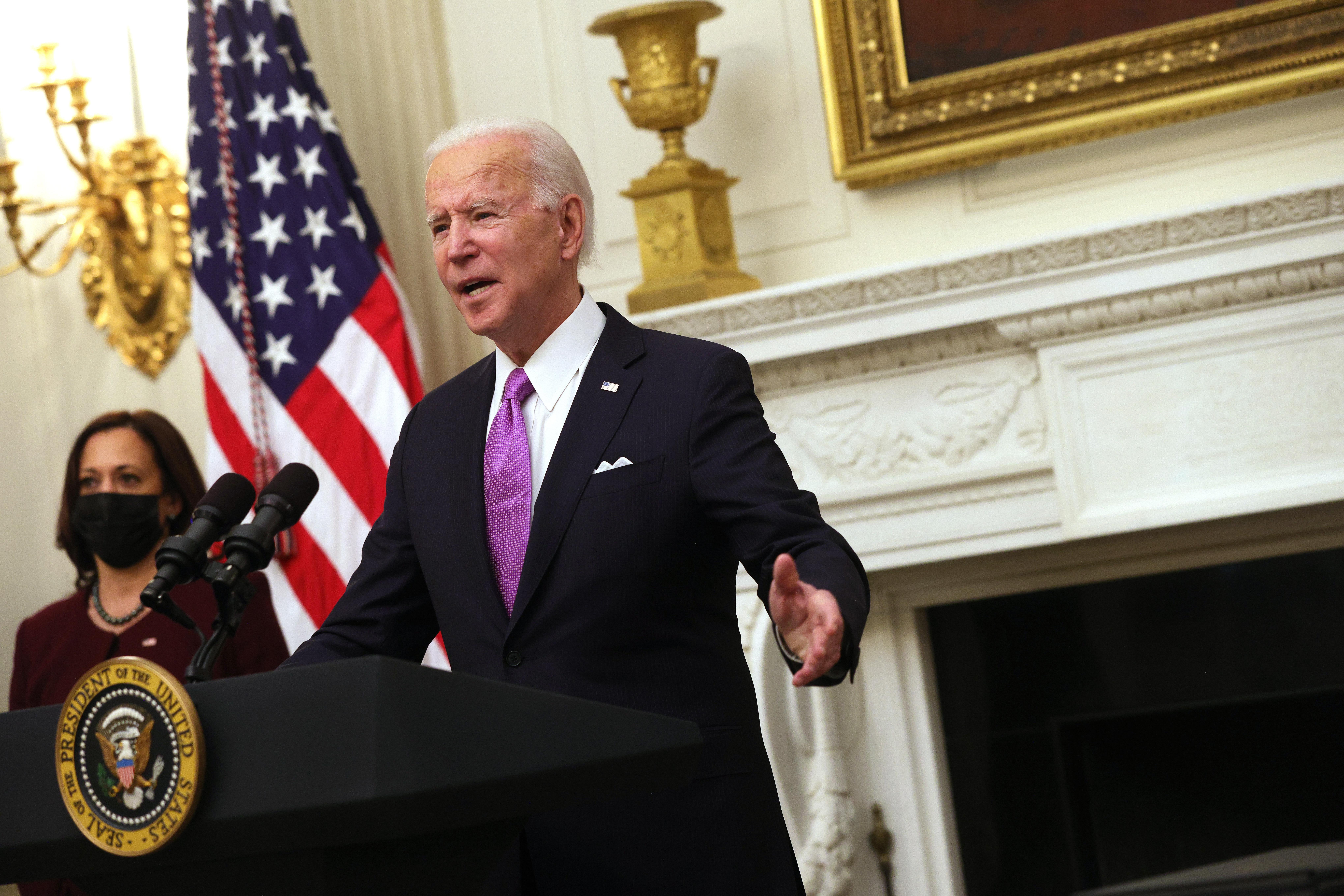 Biden raises his arms as he speaks at a podium in the State Dining Room of the White House. Vice President Kamala Harris looks on behind him.