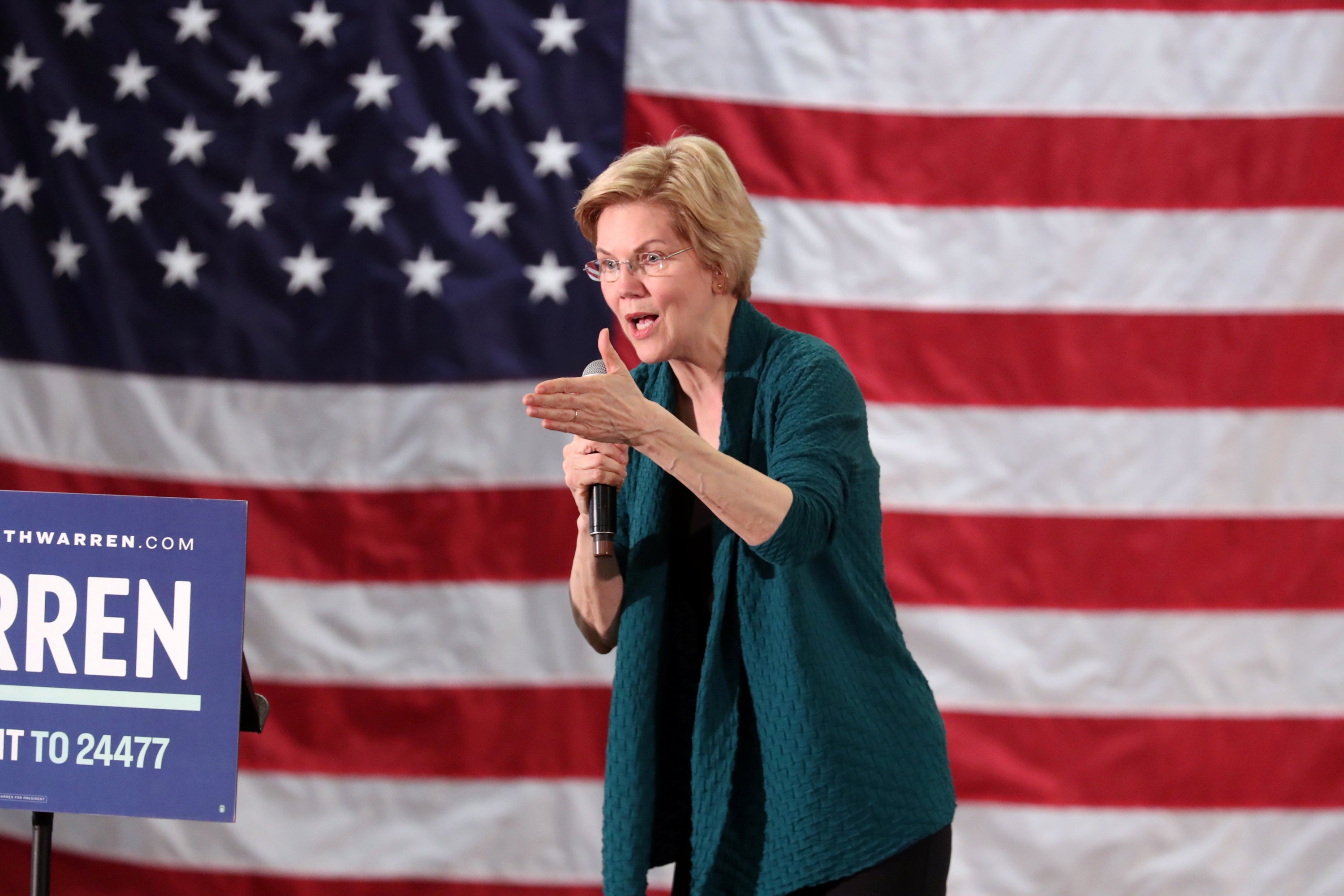 Warren, holding a microphone, speaks against an American flag backdrop.