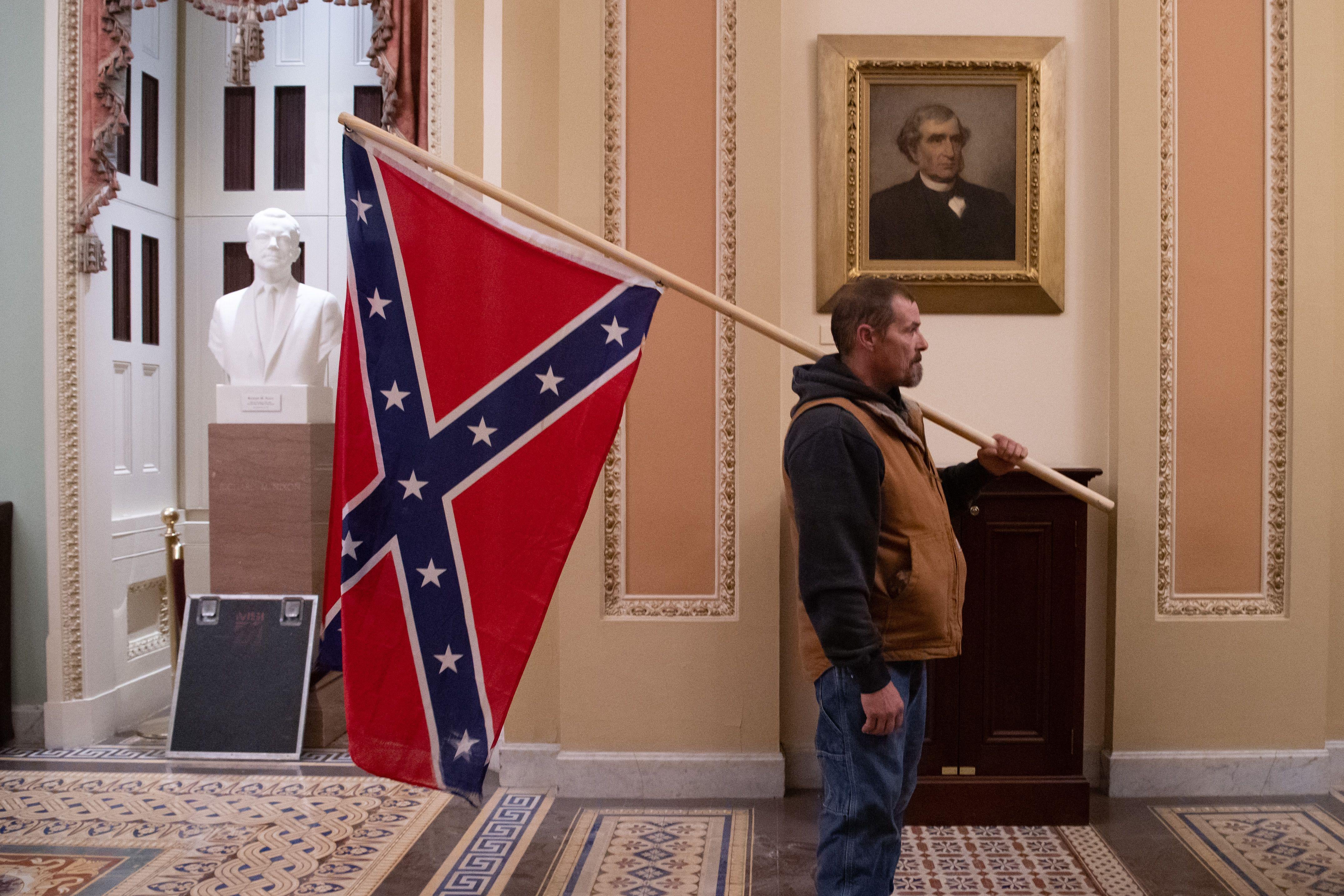 A man holding a Confederate flag stands in front of a portrait of an old member of Congress in a hallway of the Capitol.