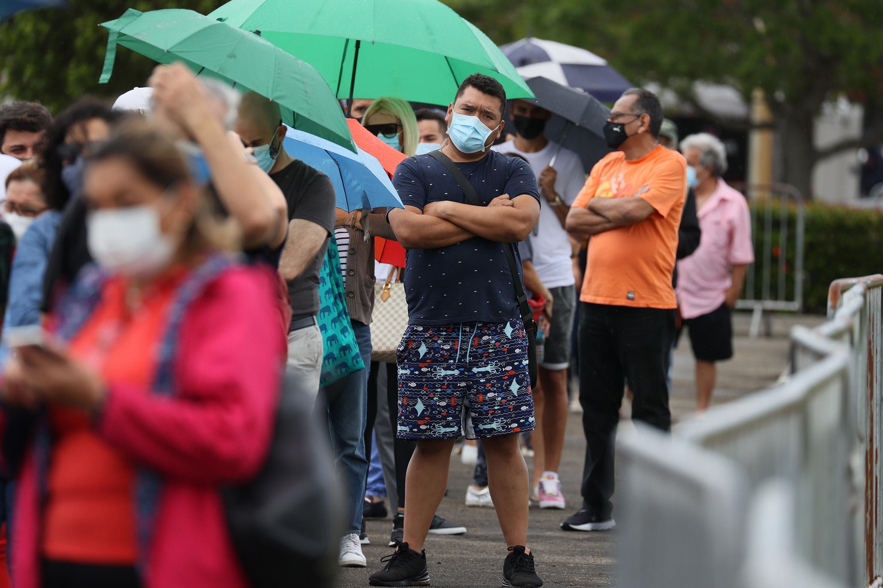 Amid a line of people holding umbrellas while waiting, a man in a mask stands with his arms crossed.