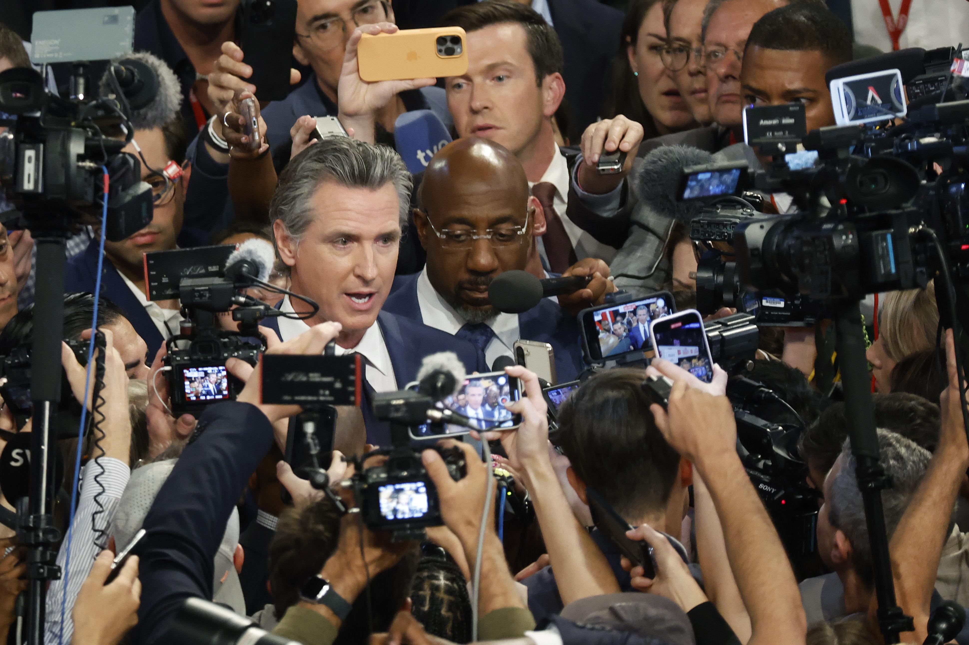 Two men speak to a swarm of reporters.