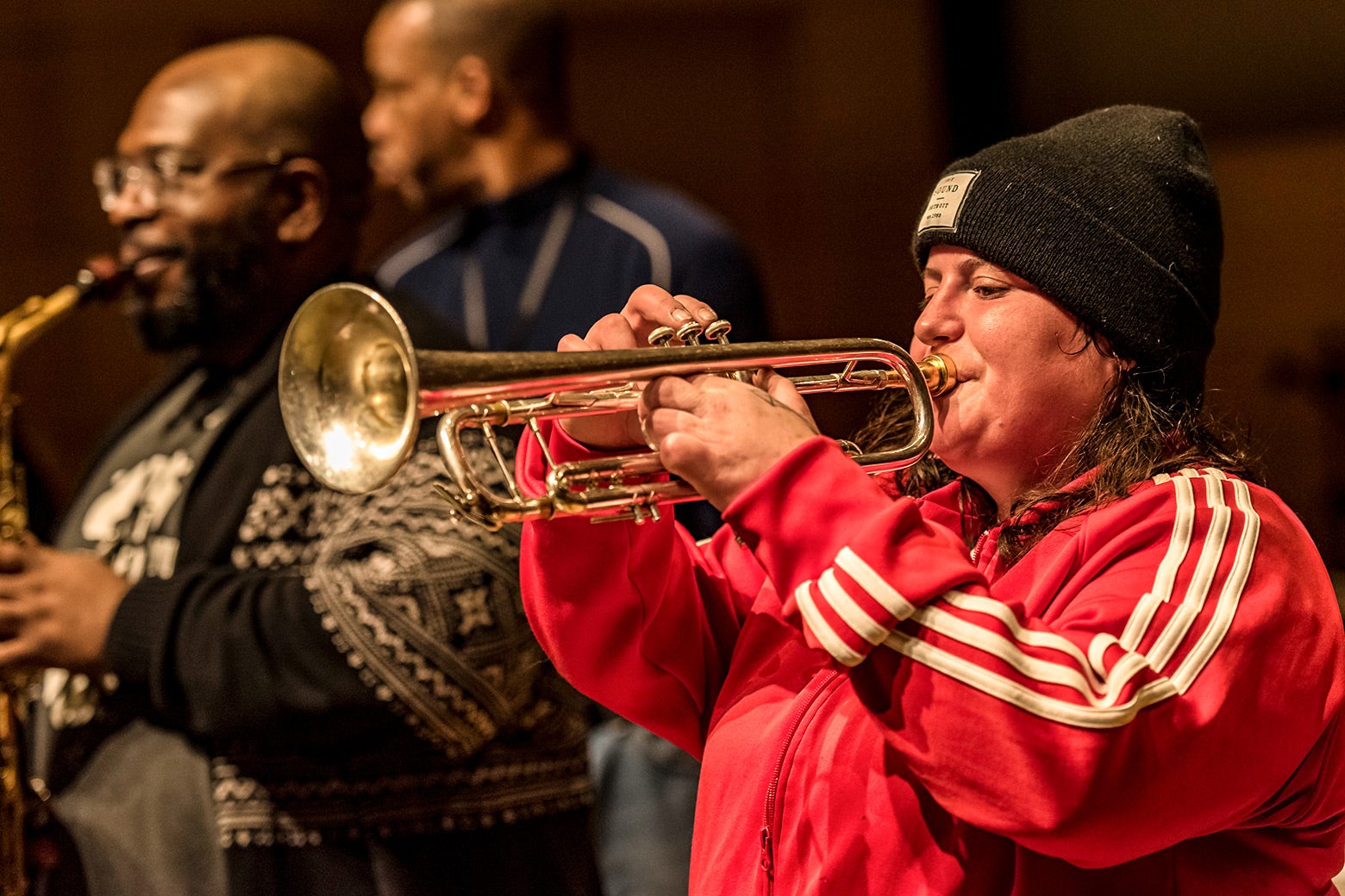 Jaimie Branch, wearing a black beanie and red Adidas tracksuit top, is seen playing trumpet with other brass musicians in the background. 