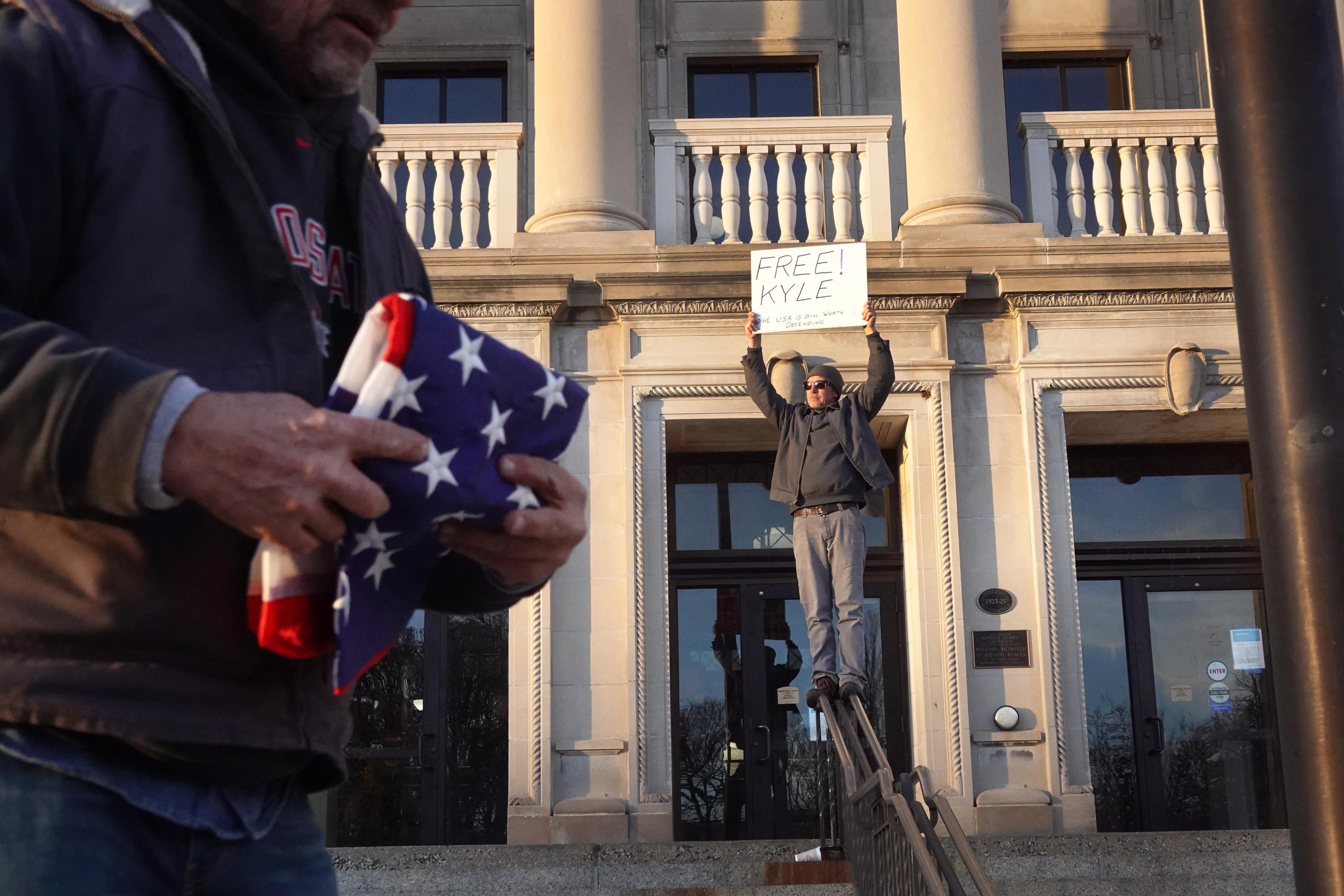 A person stands on a railing outside the courthouse holding a “Free Kyle” sign while a man in a hoodie walks in the foreground holding a folded American flag.