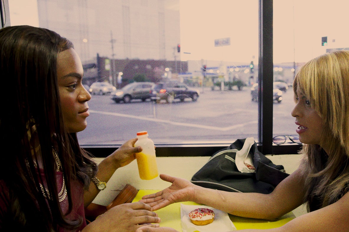 Two transgender women sit at a restaurant table talking to each other over a donut and orange juice.