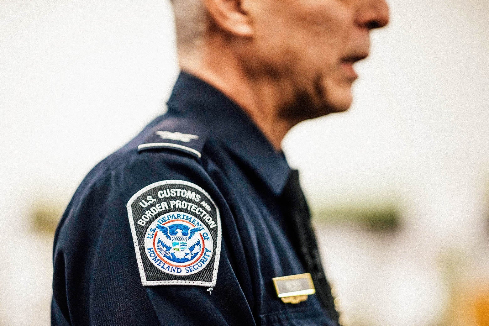 A man in a U.S. Customs and Border Protection uniform is seen from the side, with a focus on the agency's logo embroidered on his shoulder.