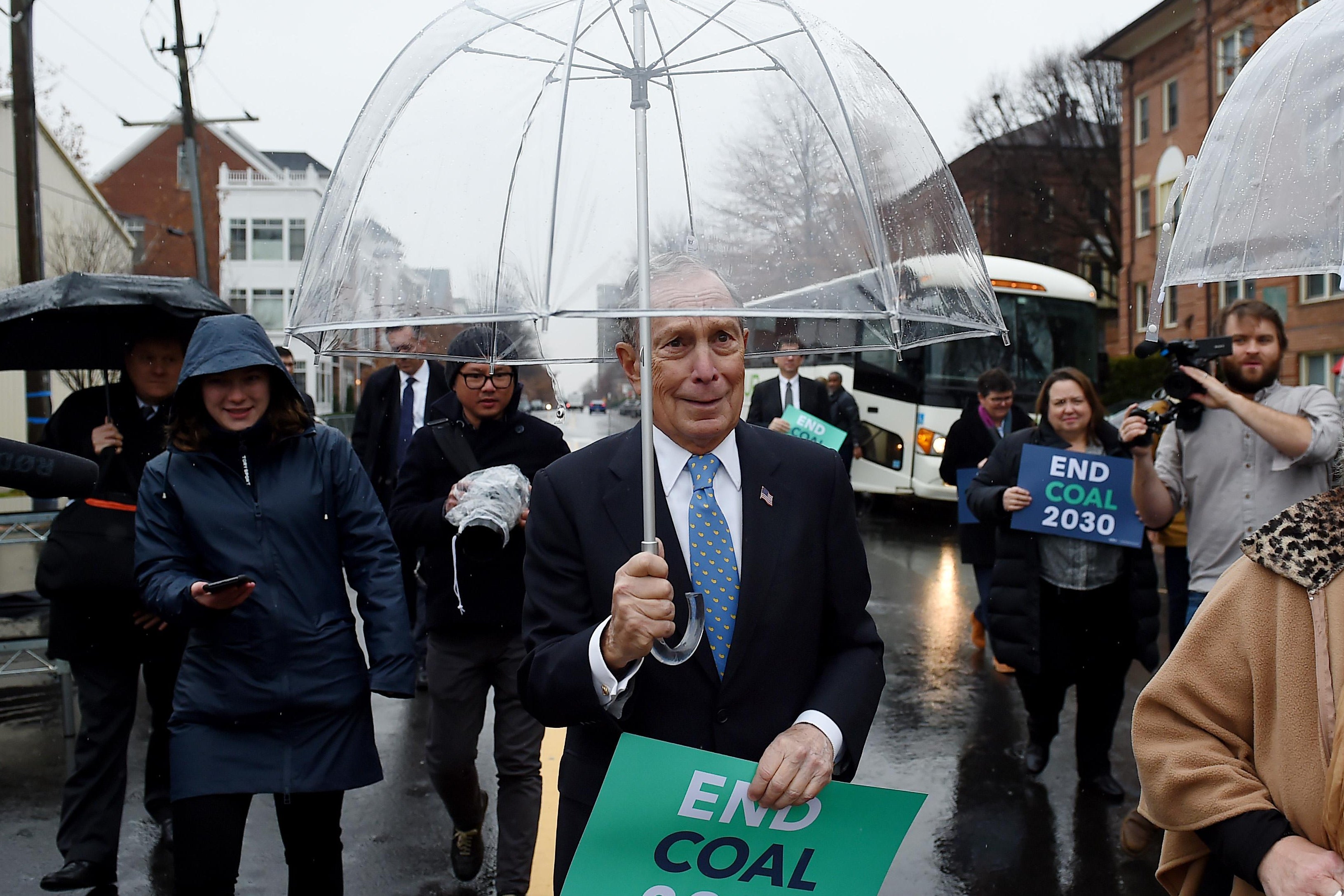 Michael Bloomberg walks with other people on a city street on a rainy day, carrying an umbrella and a sign that says "End Coal 2030."