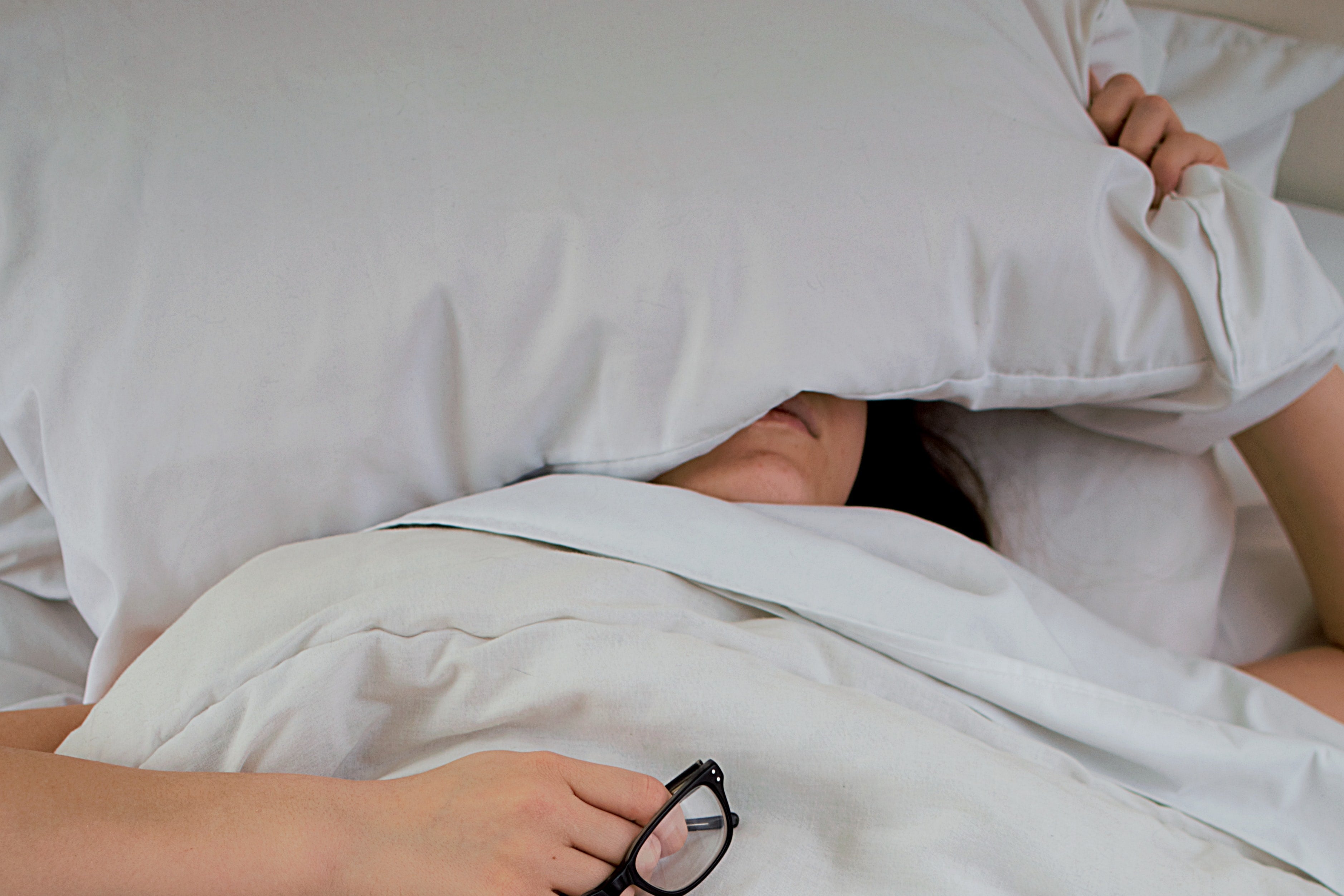A woman in bed with a pillow on top of her head and her glasses clutched in her hand.