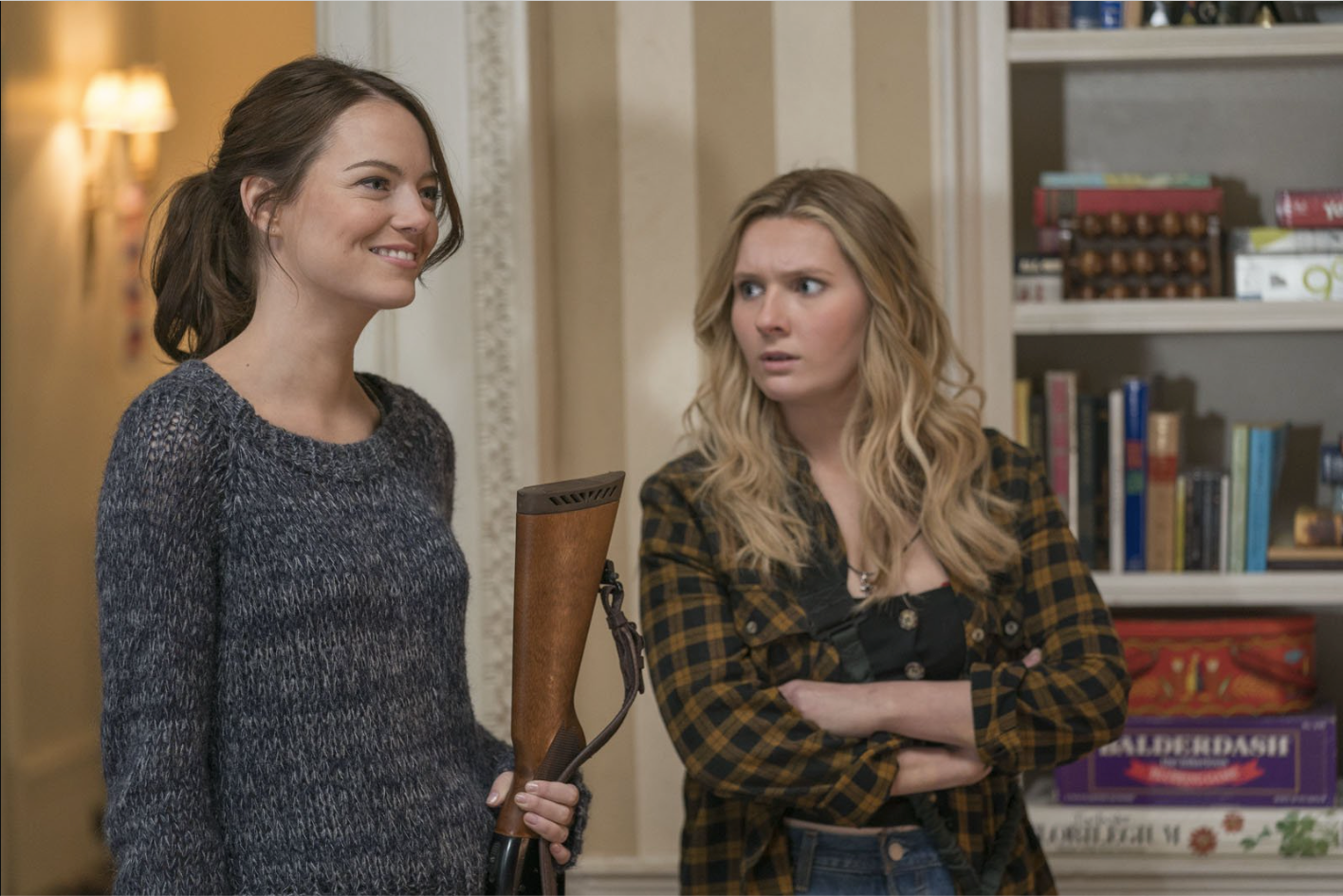 Emma Stone and Abigail Breslin stand in a room together by a bookcase. Emma Stone stands on the left in simple clothing and her hair in a ponytail, holding a rifle and smiling to something or someone off camera. Abigail Breslin stands on the right, looking at Emma Stone with her arms crossed and an incredulous look on her face. 