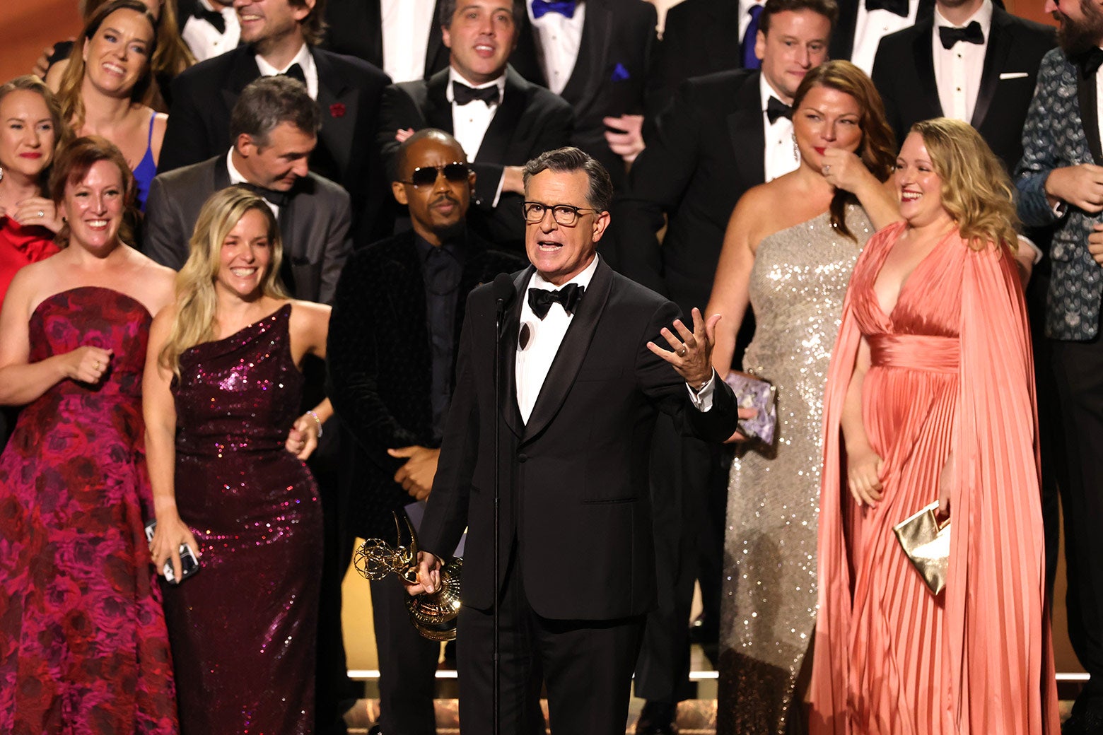 Sahnede bir Emmy ödülü tutan ve mikrofona konuşan Stephen Colbert, şovundan birçok kişiyle çevrili.