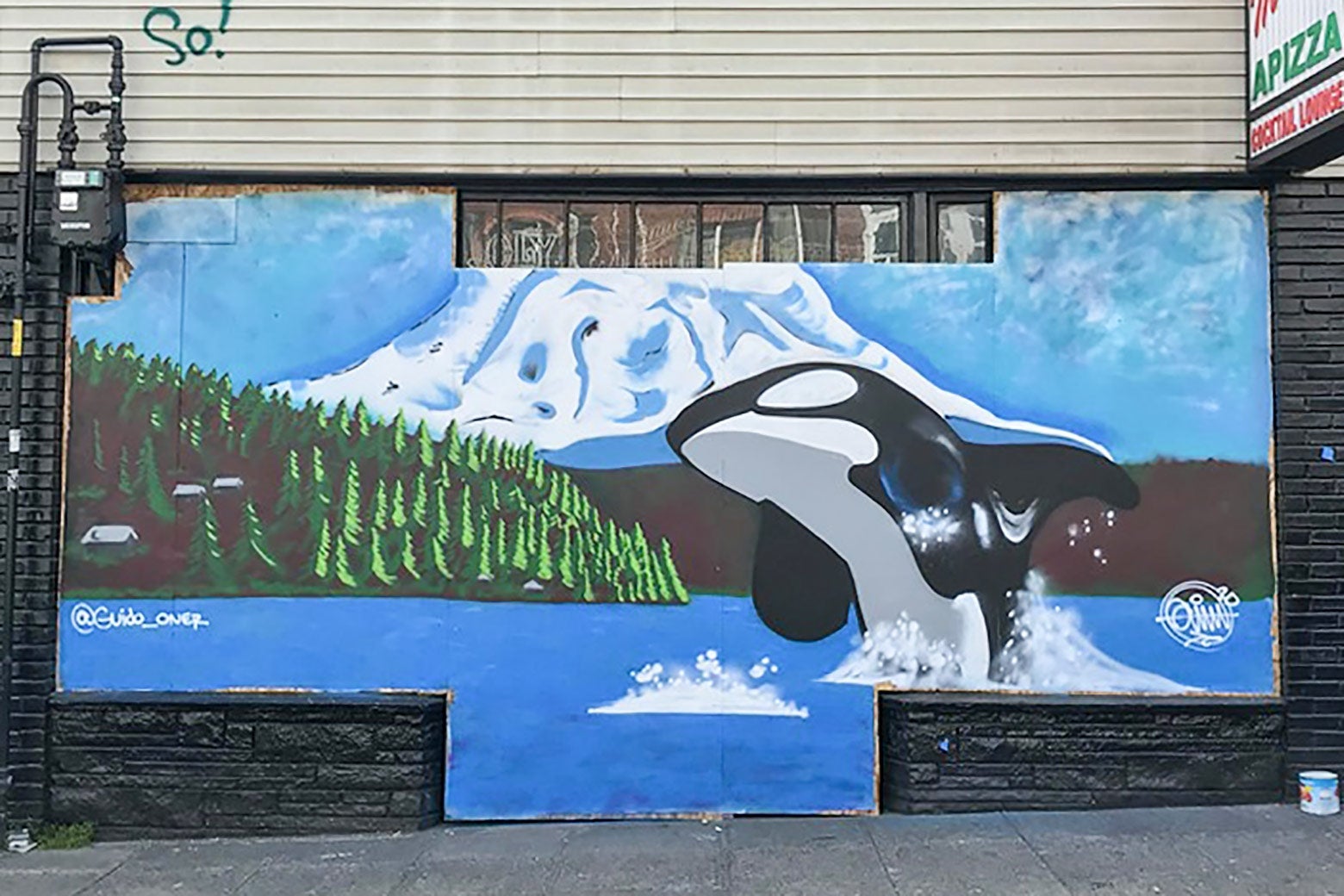 A mural beneath a sign for pizza shows an orca leaping out of a mountainside lake.
