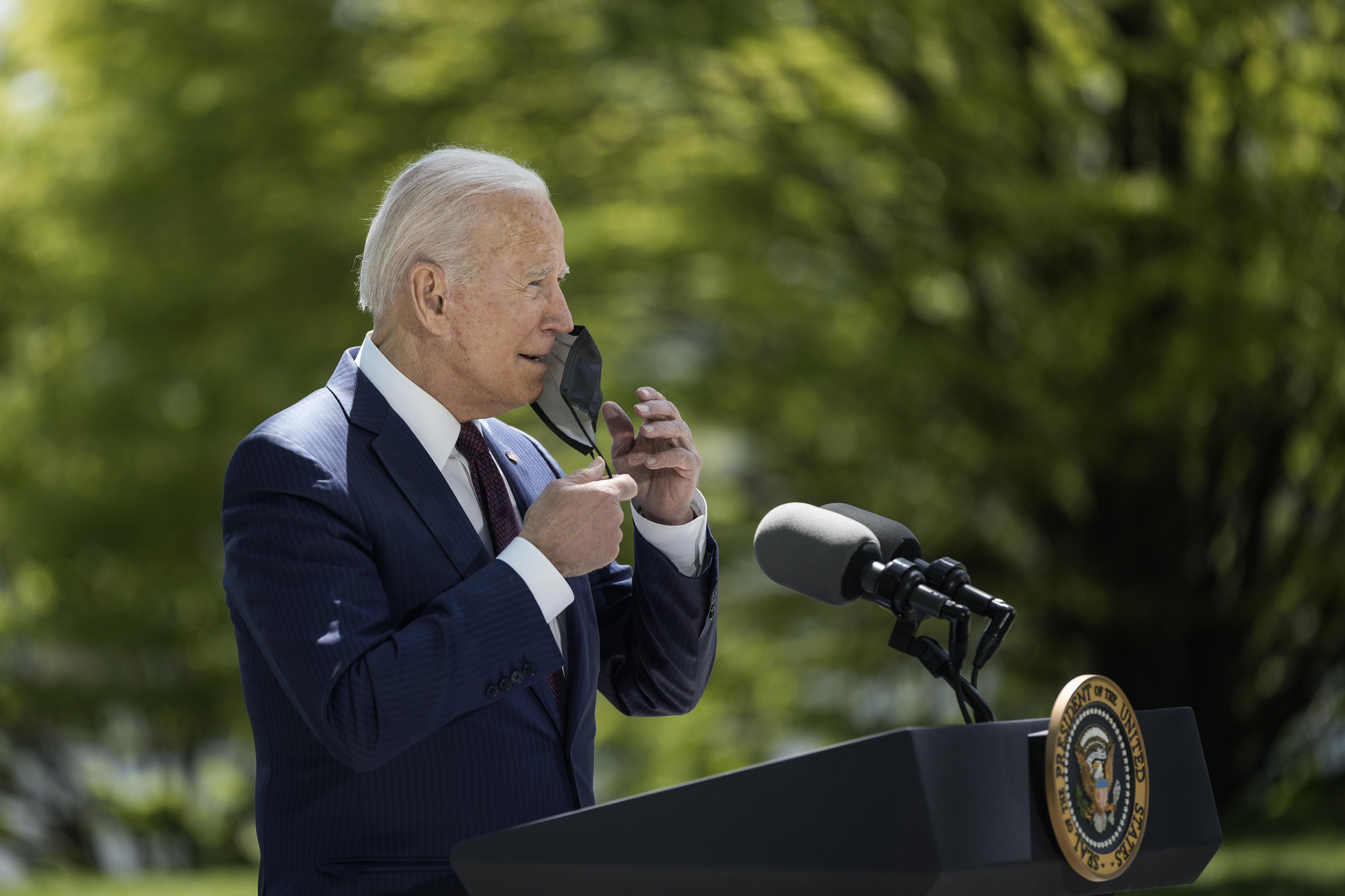Joe Biden, seen from the side, reaches up and removes a mask from his face as he stands outdoors wearing a navy suit at a presidential lectern with green trees in the background.