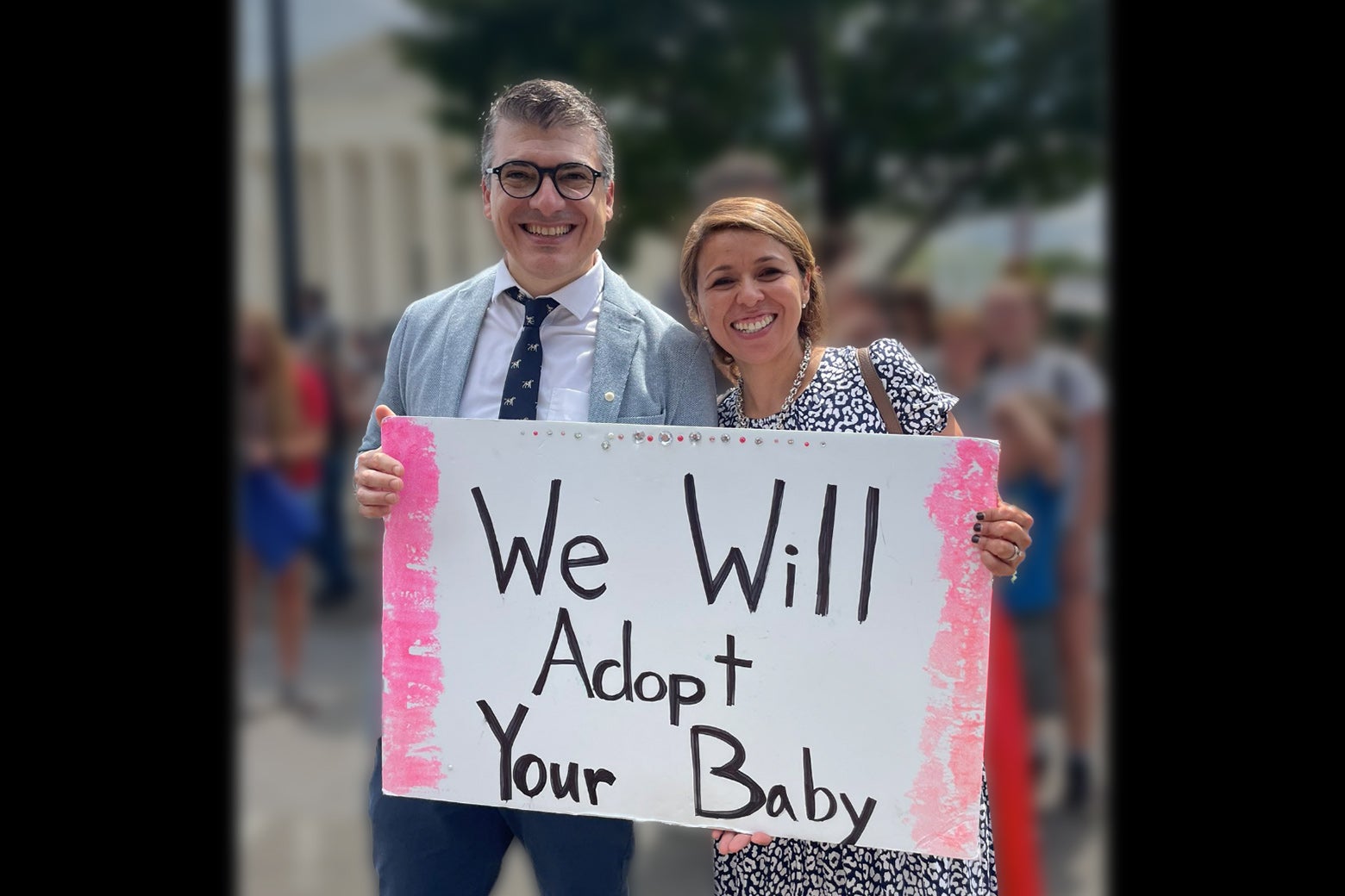 A photo of a couple holding a sign that says "We Will Adopt Your Baby" that was taken outside of the Supreme Court in Washington D.C. after Roe v Wade was overturned.