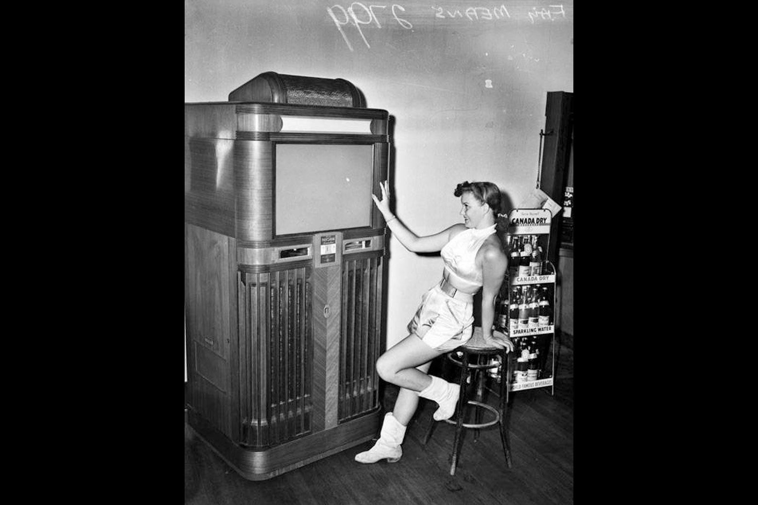 A black-and-white image of a white woman in 1940s getup sitting in front of a wood Panoram, touching the screen admiringly and watching. 