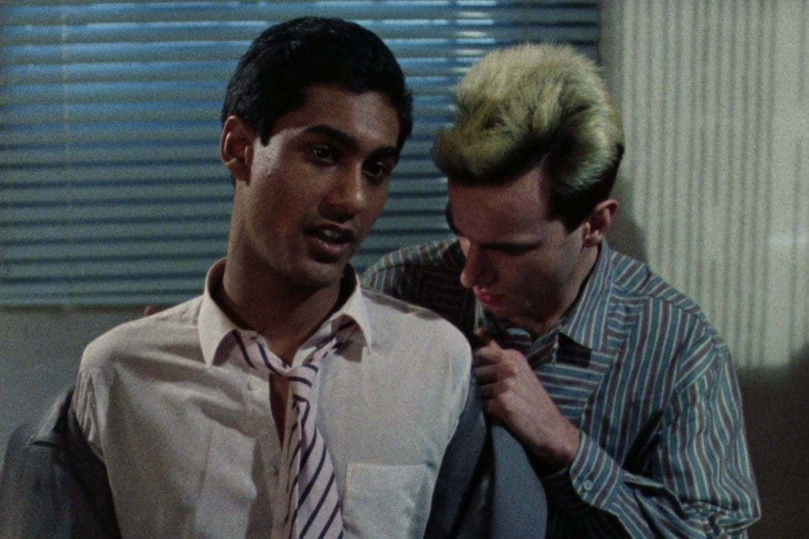 A young blonde white man in a blue and white striped button down nearly buries his head in the crook of a young South Asian man's neck as he helps the other man take off his suit jacket. 