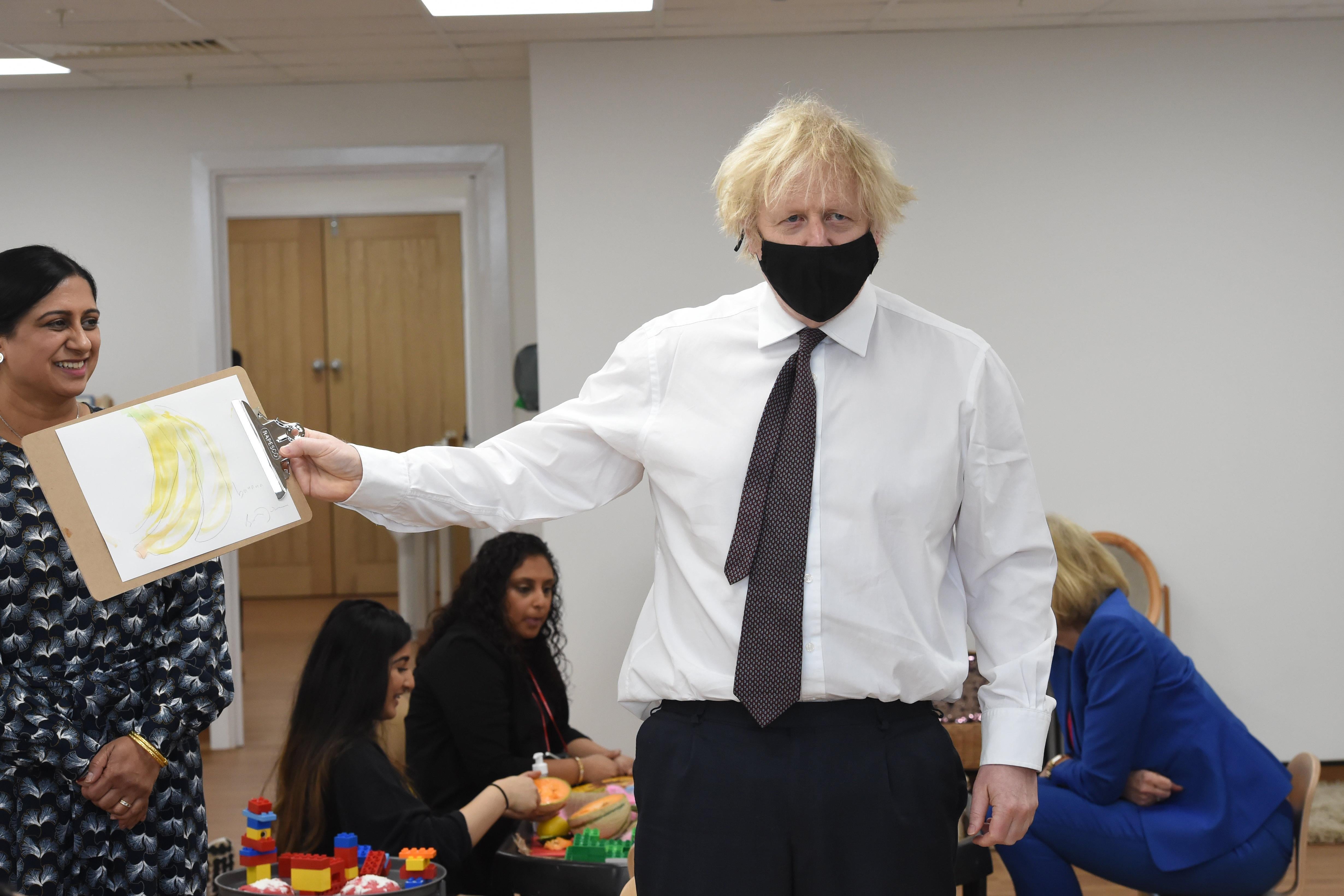 Boris Johnson stands in a classroom and holds up a clipboard with a drawing of a bunch of bananas.