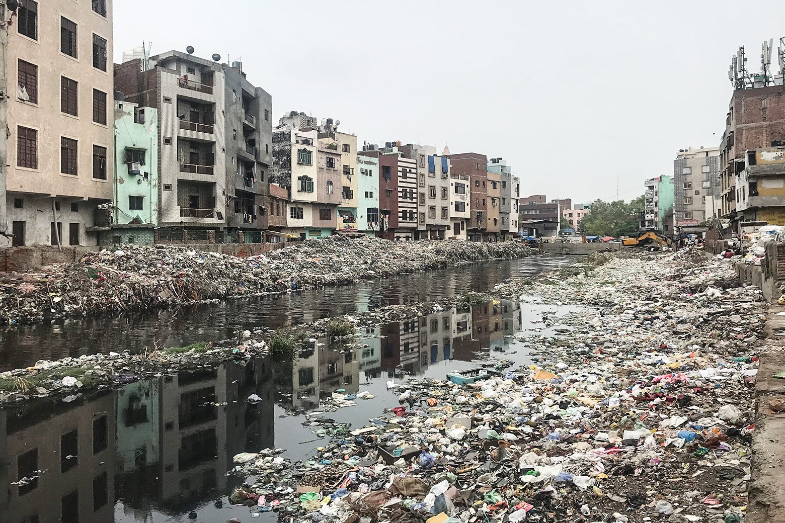 A drainage canal with murky water and filled with trash. On both sides, there are trash-filled banks and five- or six-story buildings.
