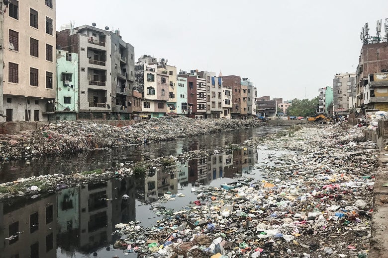 A drainage canal with murky water and filled with trash. On both sides, there are trash-filled banks and five- or six-story buildings. 