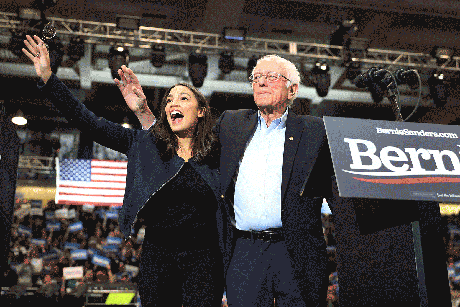 AOC and Bernie Sanders stand next to each other onstage in an arena and wave to the crowd as an illustrated version of Biden sidles up beside them.