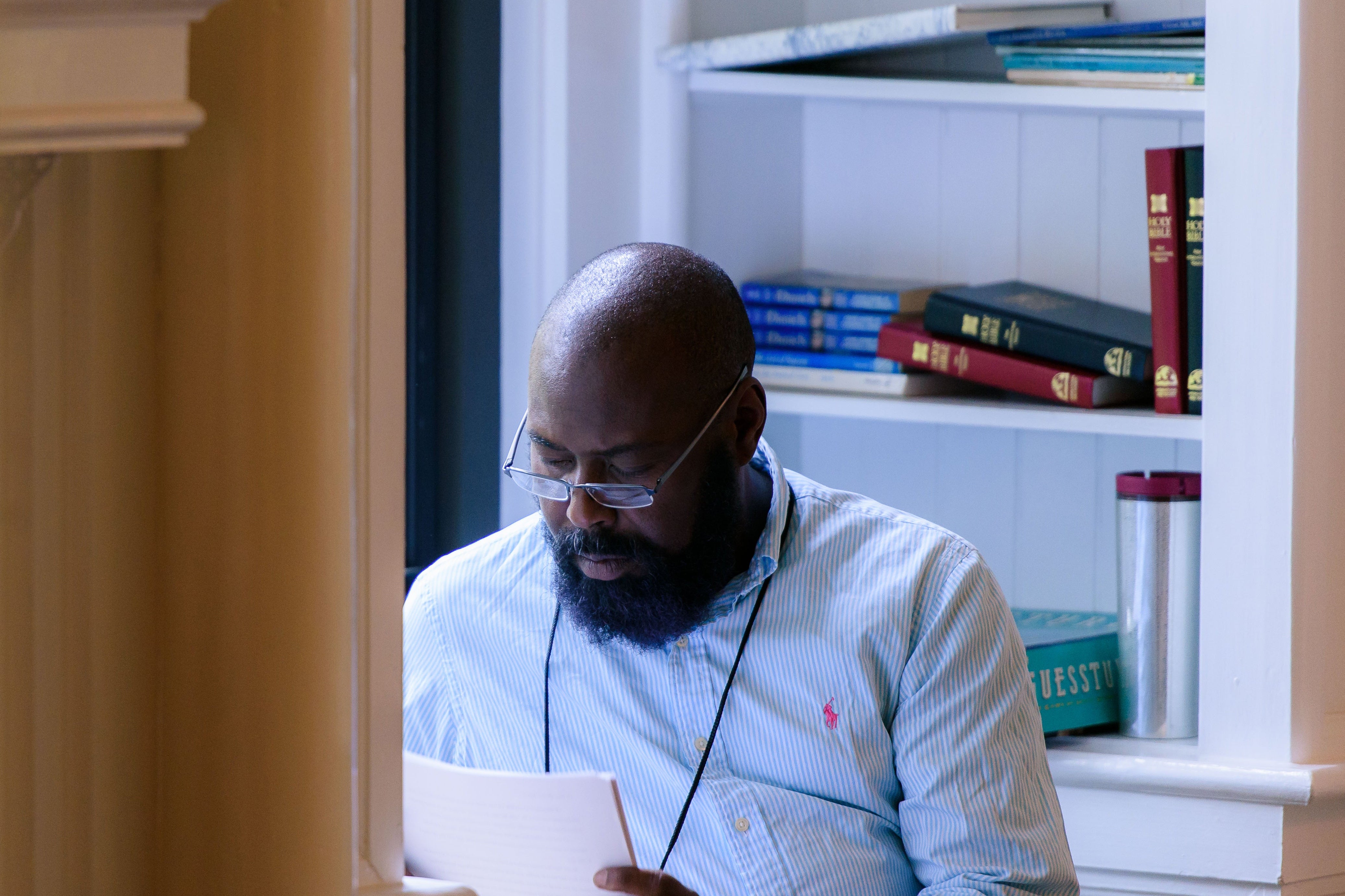 Randall Kenan wears glasses and holds a sheaf of papers in front of shelves of books.