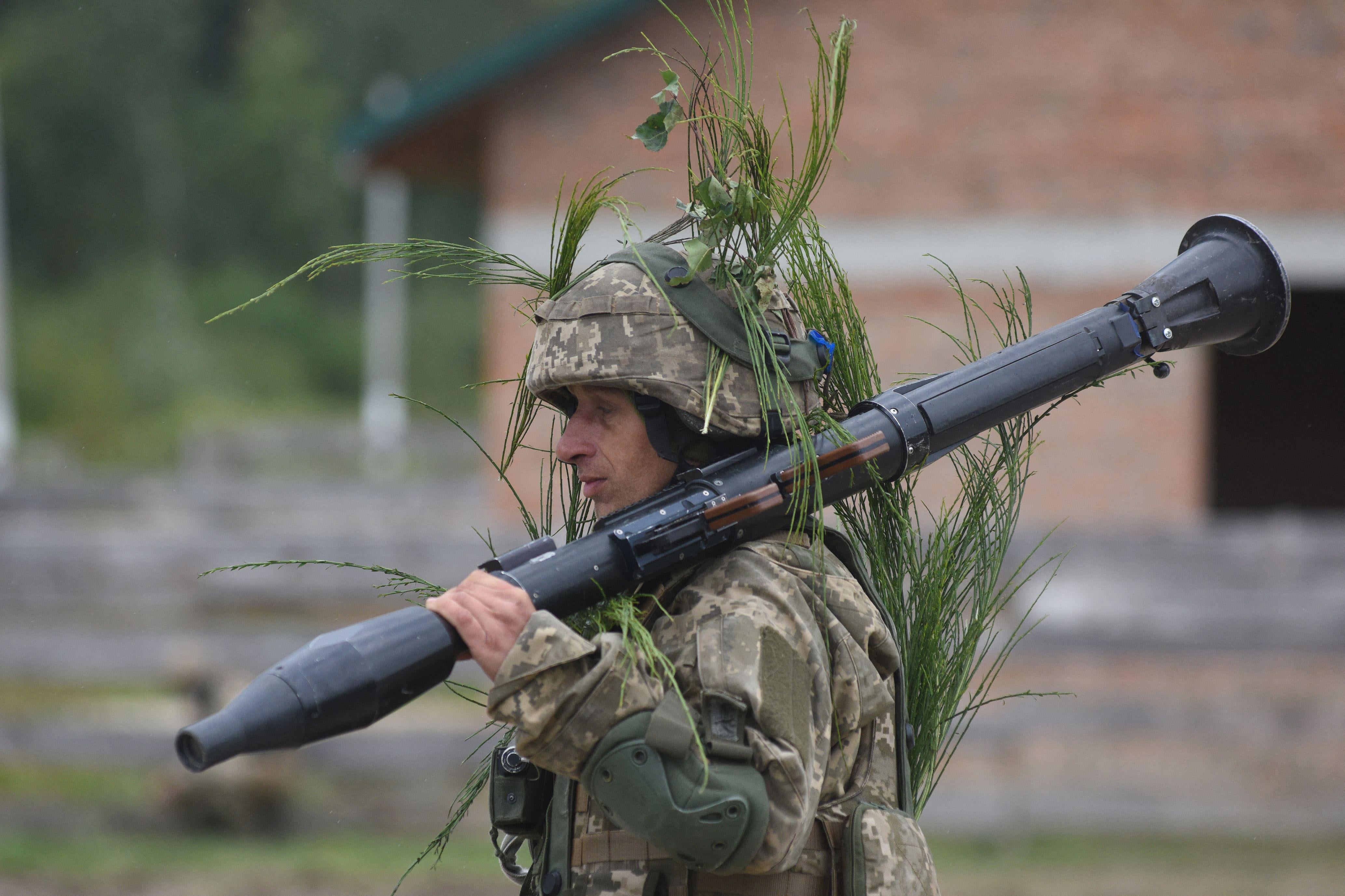 A soldier in fatigues walks outside holding a bazooka over his shoulder
