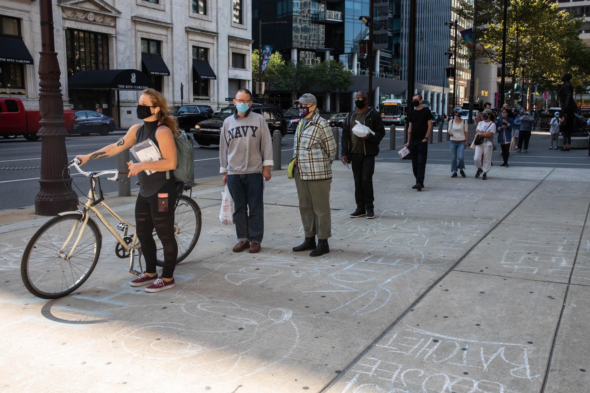 People wearing masks wait in a socially distanced line on the sidewalk