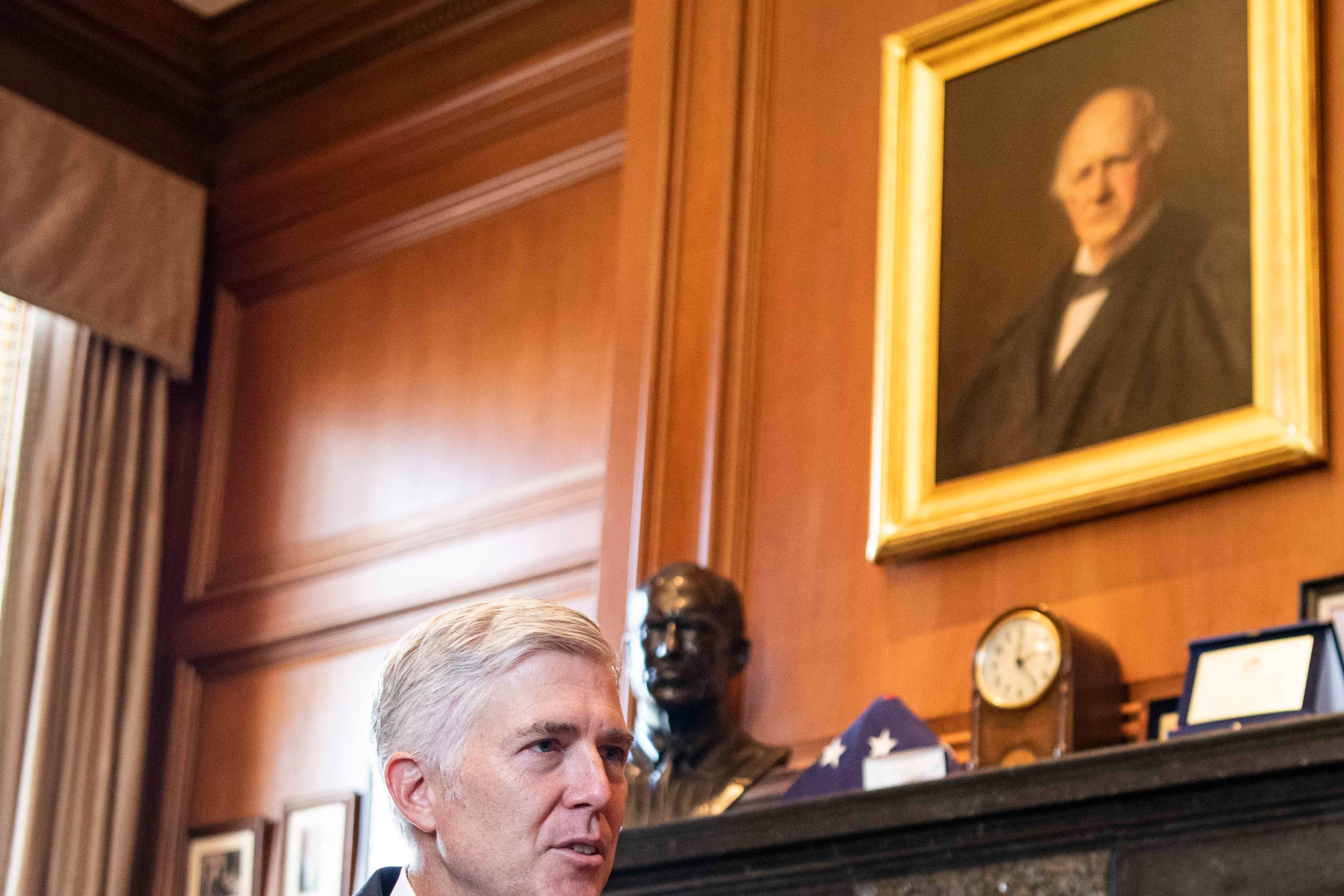 Neil Gorsuch, seated in front of a fireplace, talking with his hands held up.