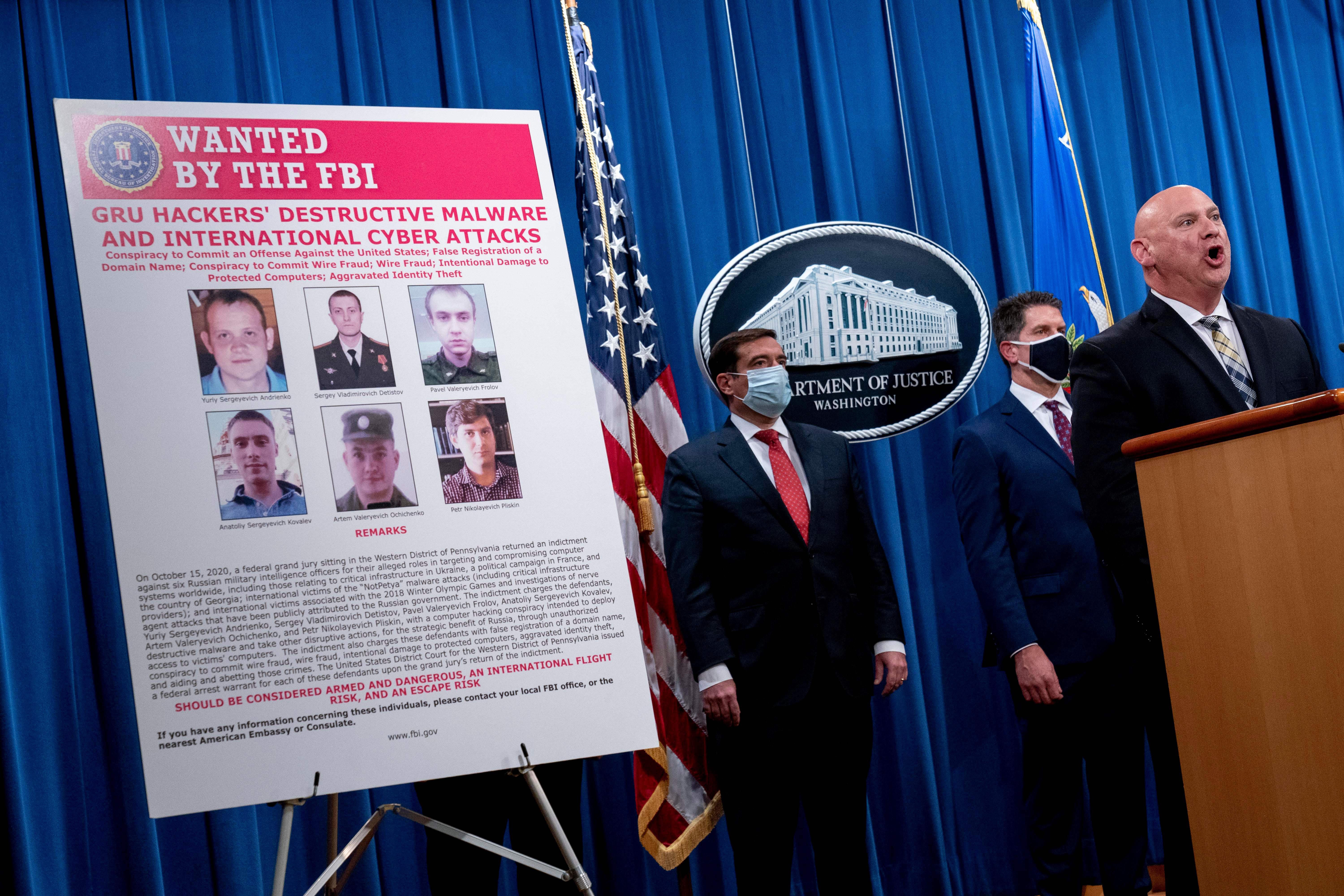 A man speaks at a lectern in front of two men wearing masks and a Wanted poster showing six men wanted by the FBI. 