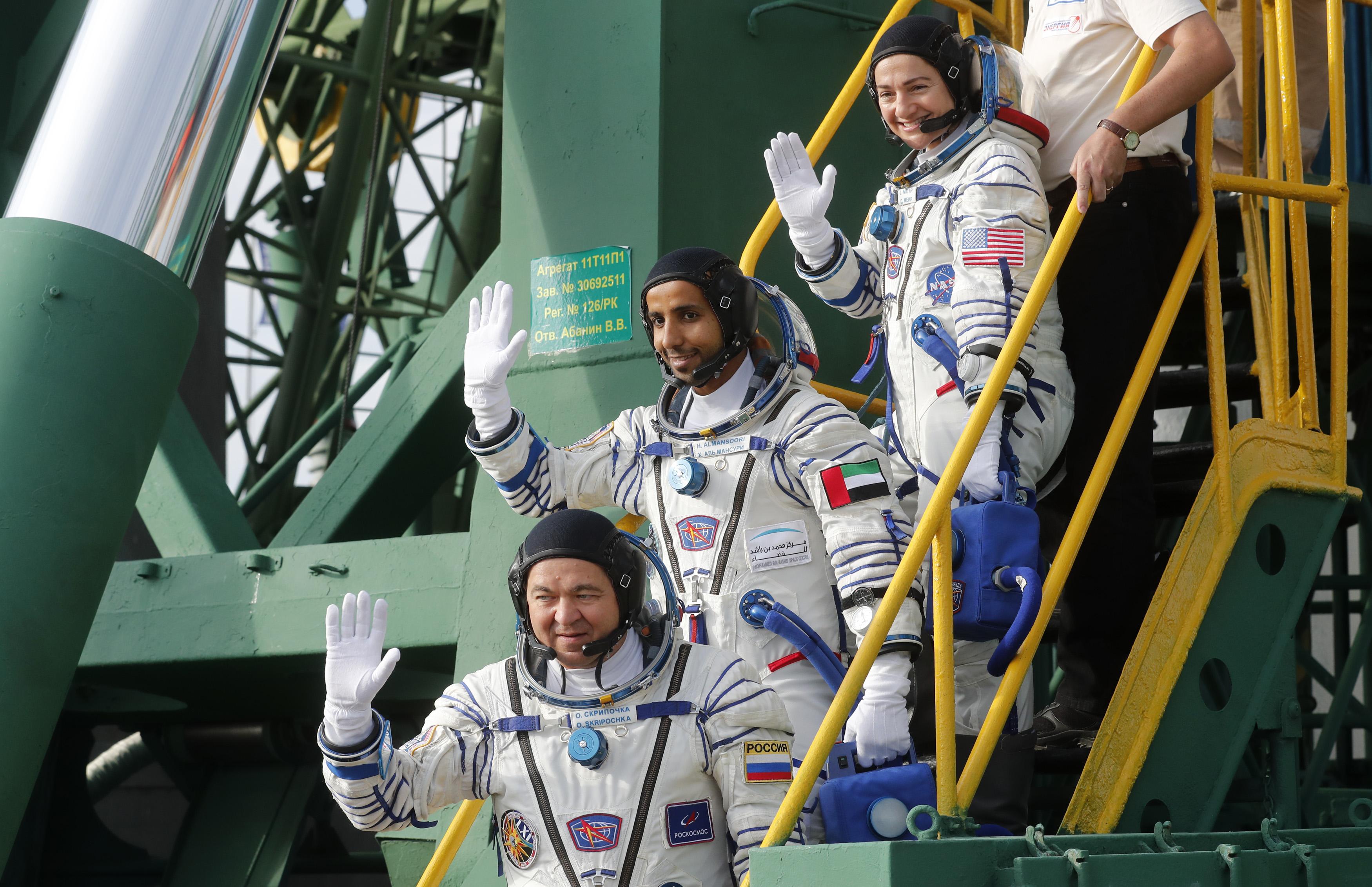 Three astronauts walk down stairs, dressed in space suits with the Russian, U.S. and UAE flags on their sleeves. Behind and surrounding them is a metal structure for a rocket launch. 