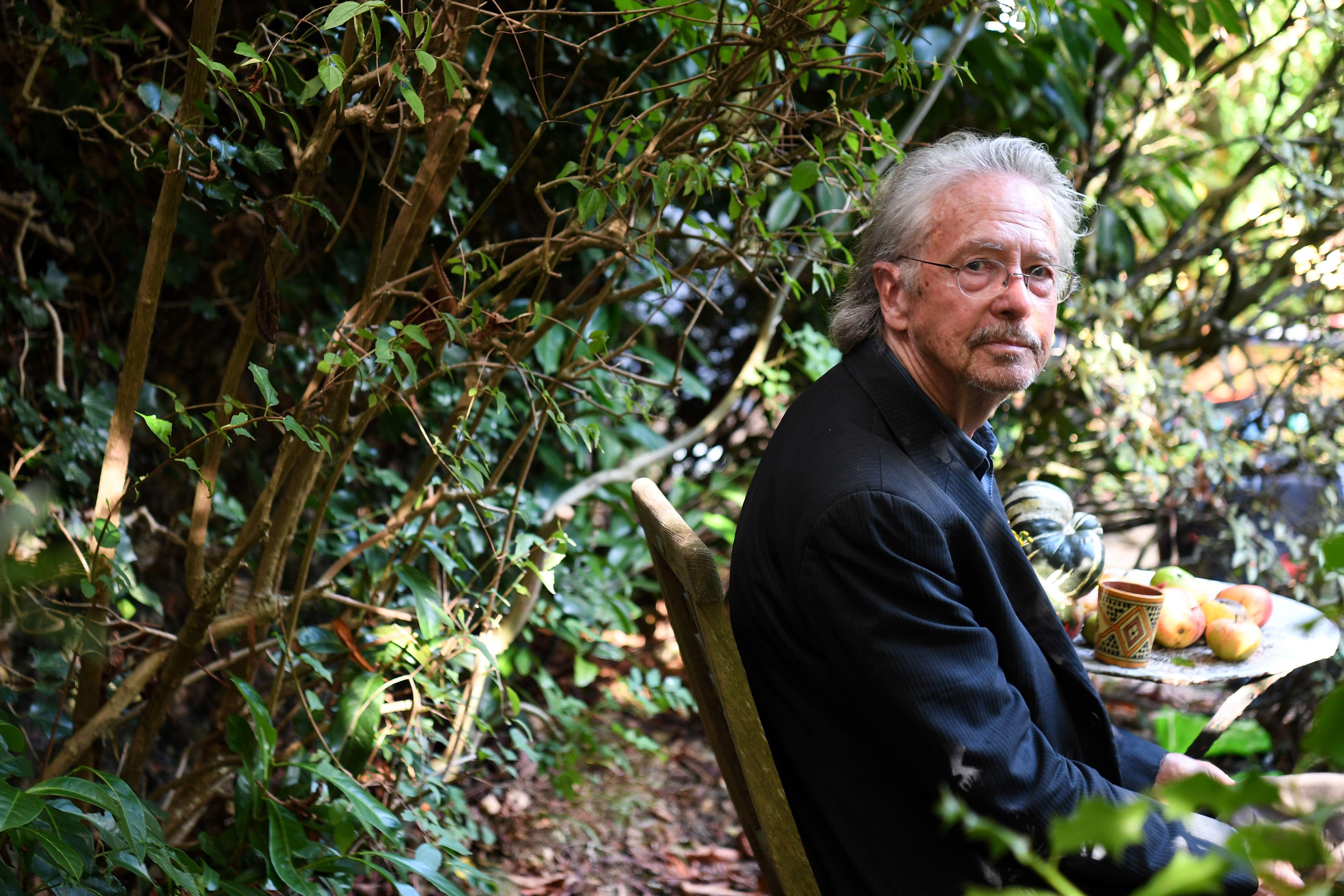 Peter Handke sits at a table with a mug and several apples against a backdrop of greenery.