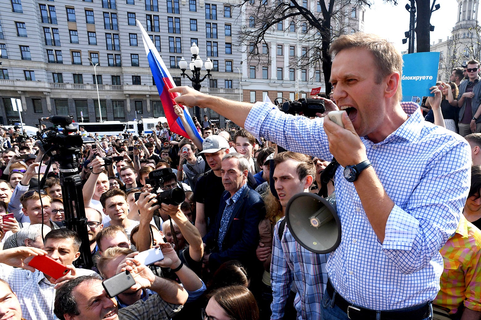 Navalny speaks into a speaker, attached to a megaphone, and points, to a crowd of protesters, a Russian flag hoisted in the crowd.