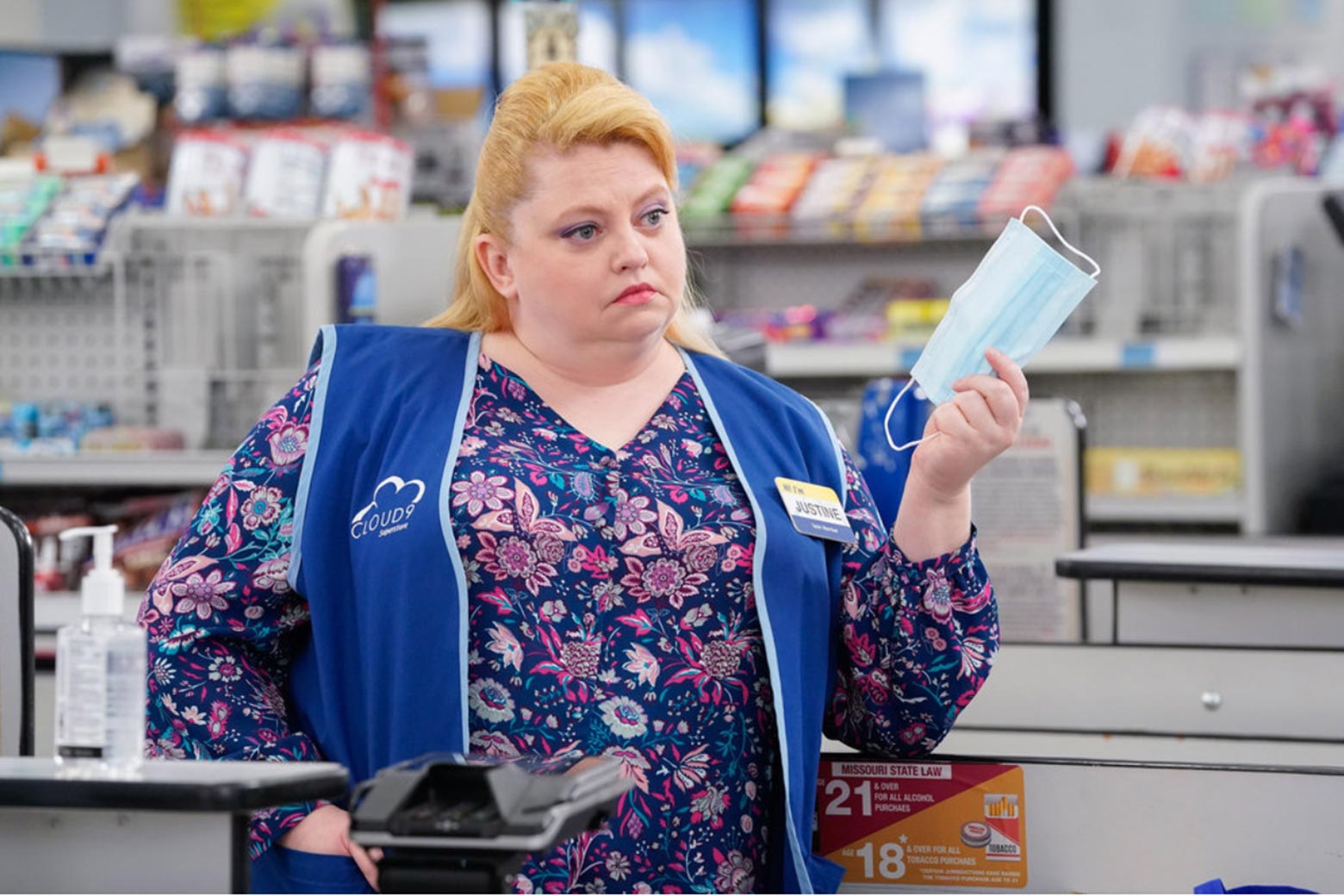 a woman stands at a cash register in a big box store, skeptically holding a surgical mask