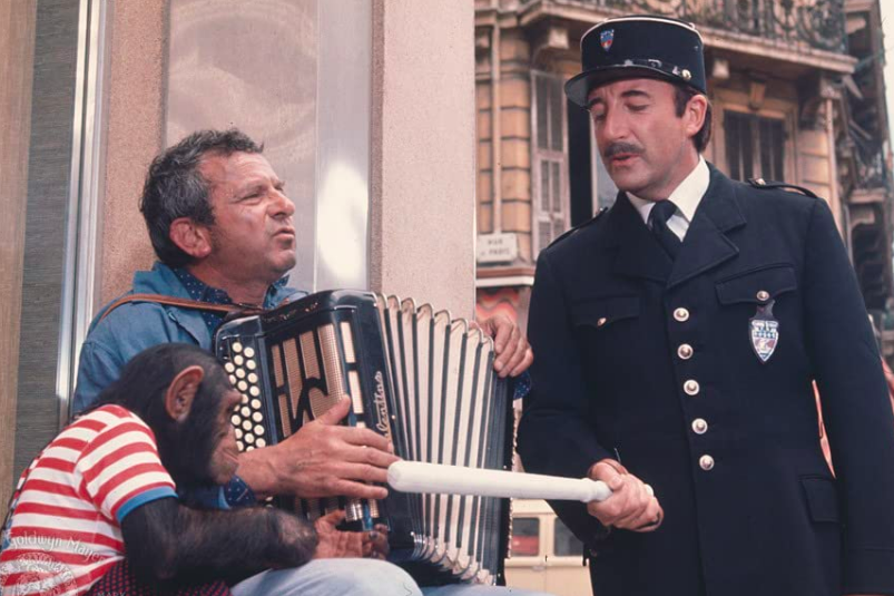 Peter Sellers, dressed in a police uniform, points a baton at a monkey wearing a striped shirt. John Bluthal plays an accordian.