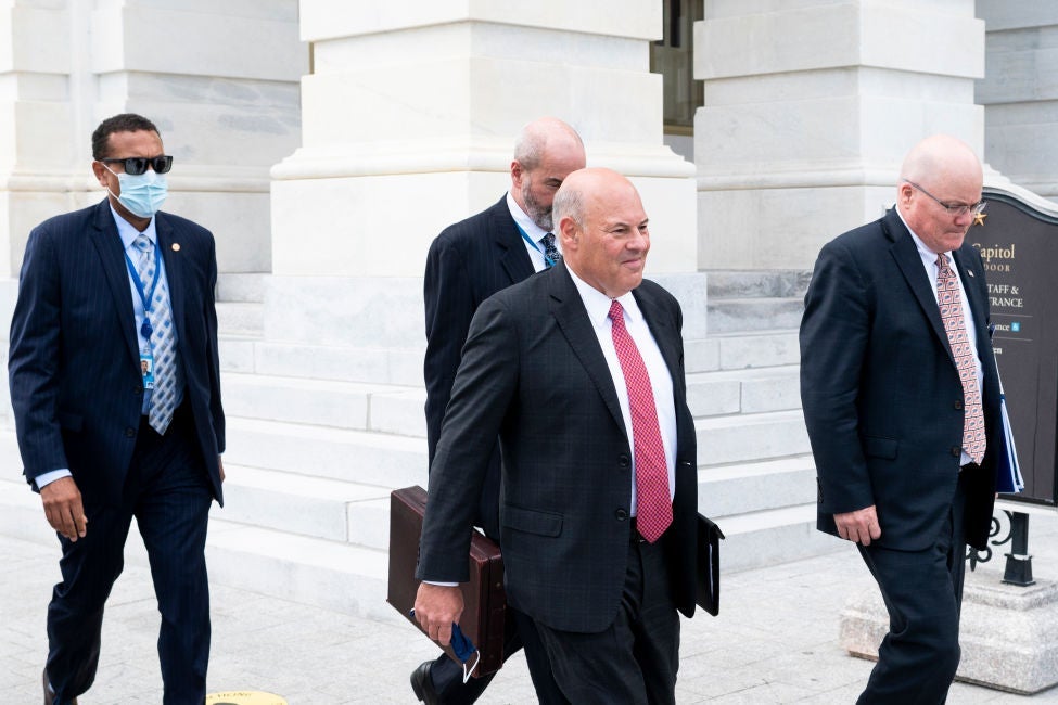 DeJoy and three other men wearing dark suits walk past steps and columns on the exterior of the Capitol building.