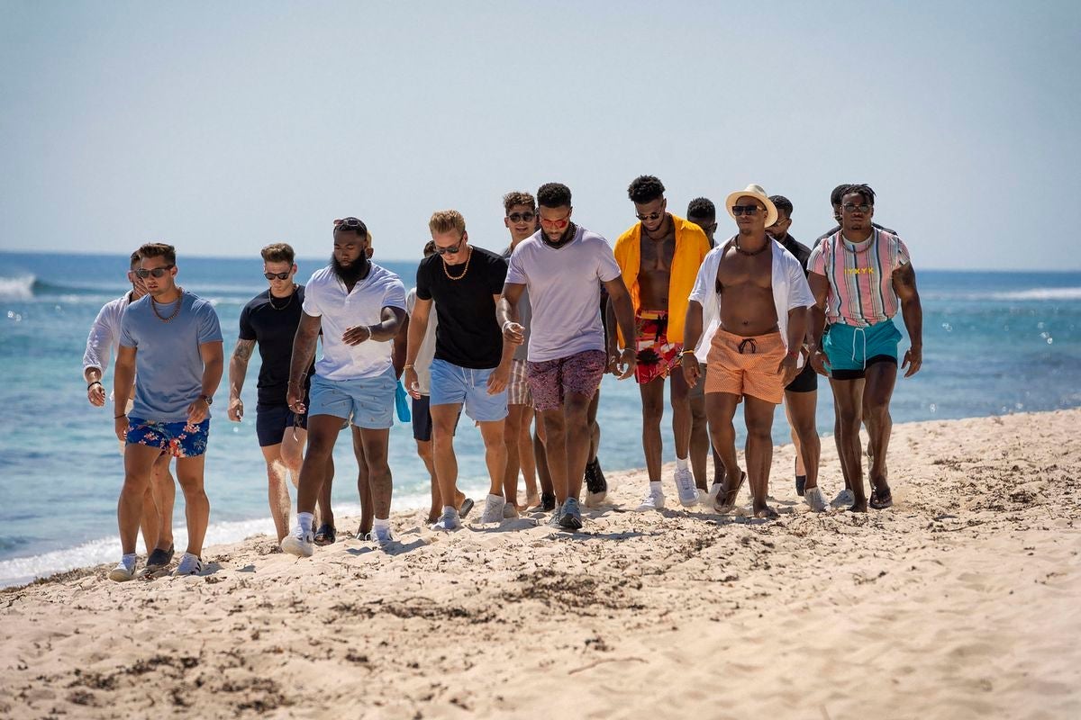 A multiracial group of a dozen men, trudging along a sandy beach next to the water, all wearing short sleeved shirts and sunglasses.
