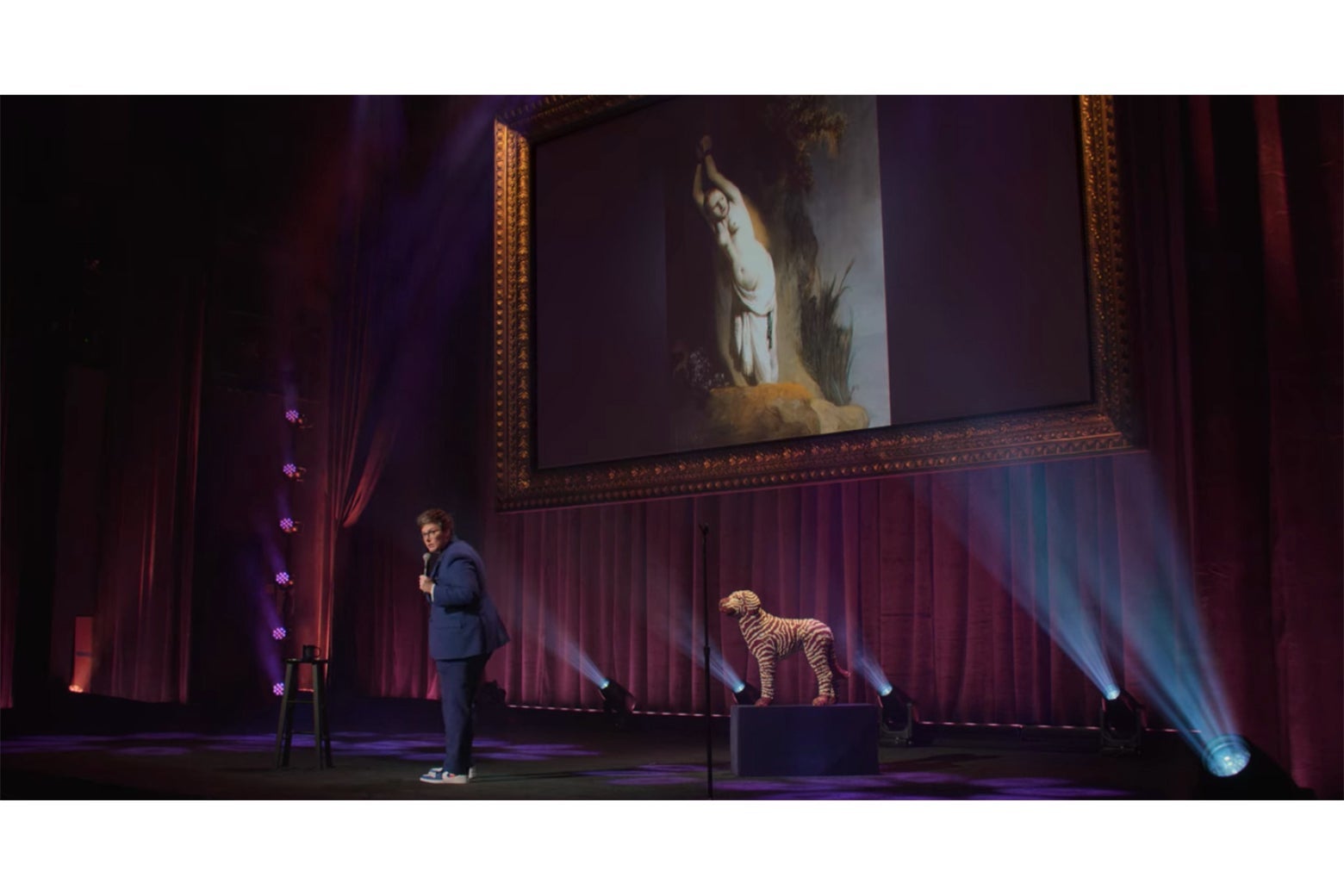 Hannah Gadsby onstage in front of a Rembrandt painting of Andromeda chained to her rock.