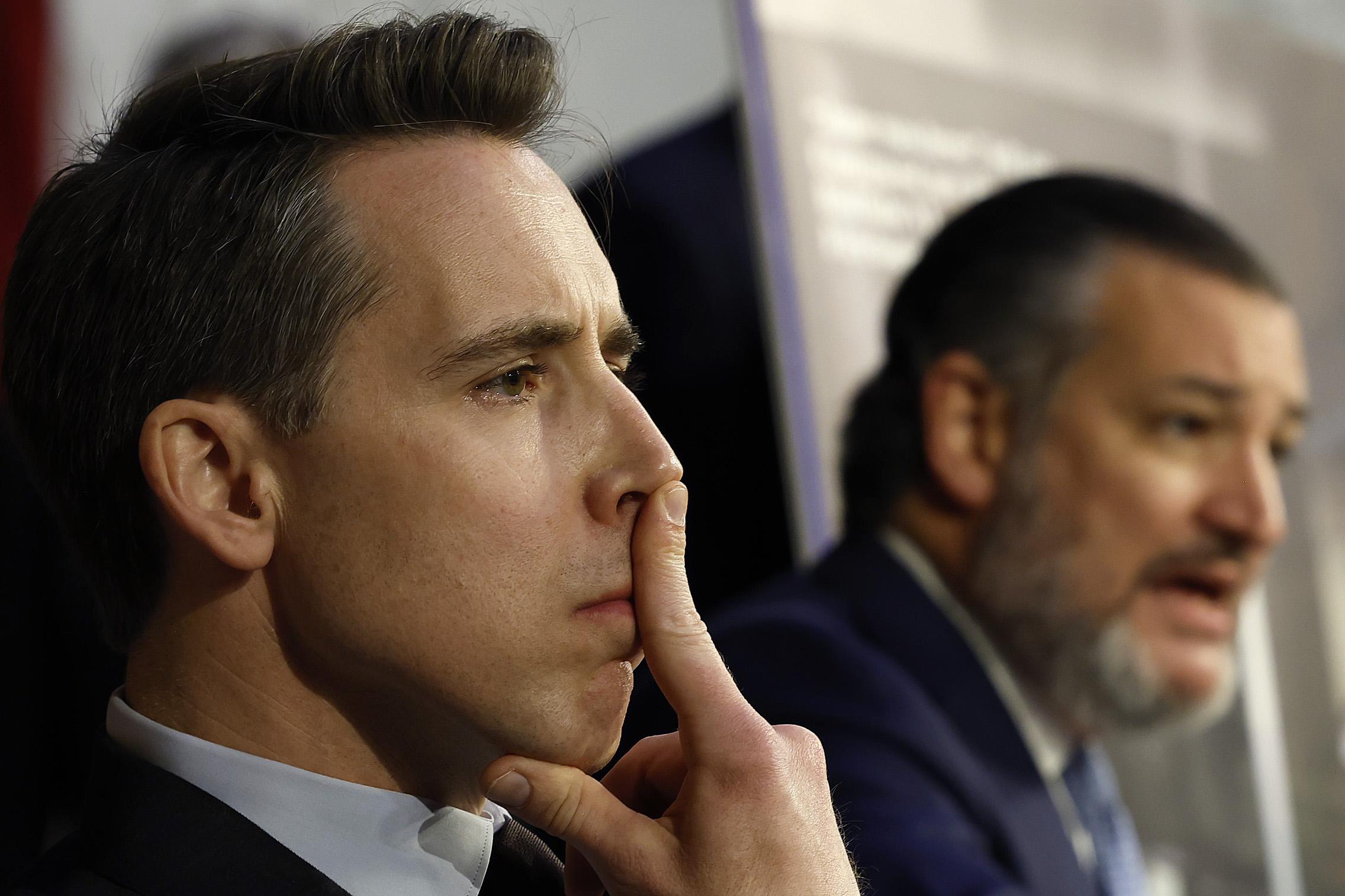 Hawley, whose profile is in the foreground, presses a finger to his nose and lip, in an expression of concentration. Behind him, out of focus, Ted Cruz appears to be speaking.  