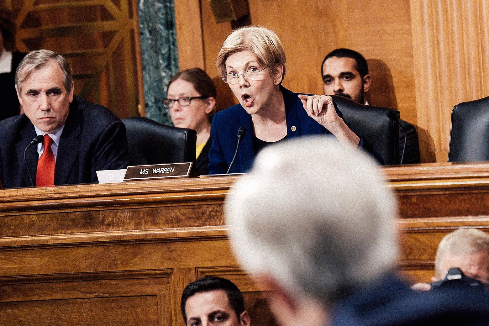 Elizabeth Warren, sitting at a panel while surrounded by others, speaks into a microphone.