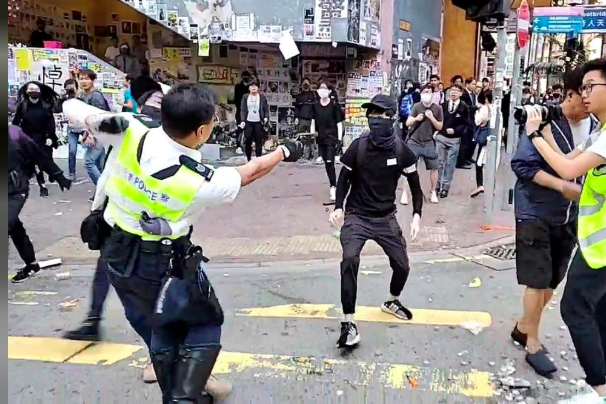 A police officer aims his gun at a black-clad protester as others look on.