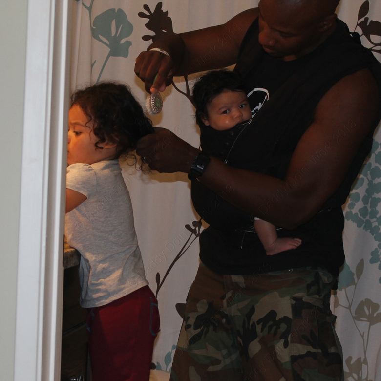 The author wearing his baby in a chest carrier while he brushes his older daughter’s hair