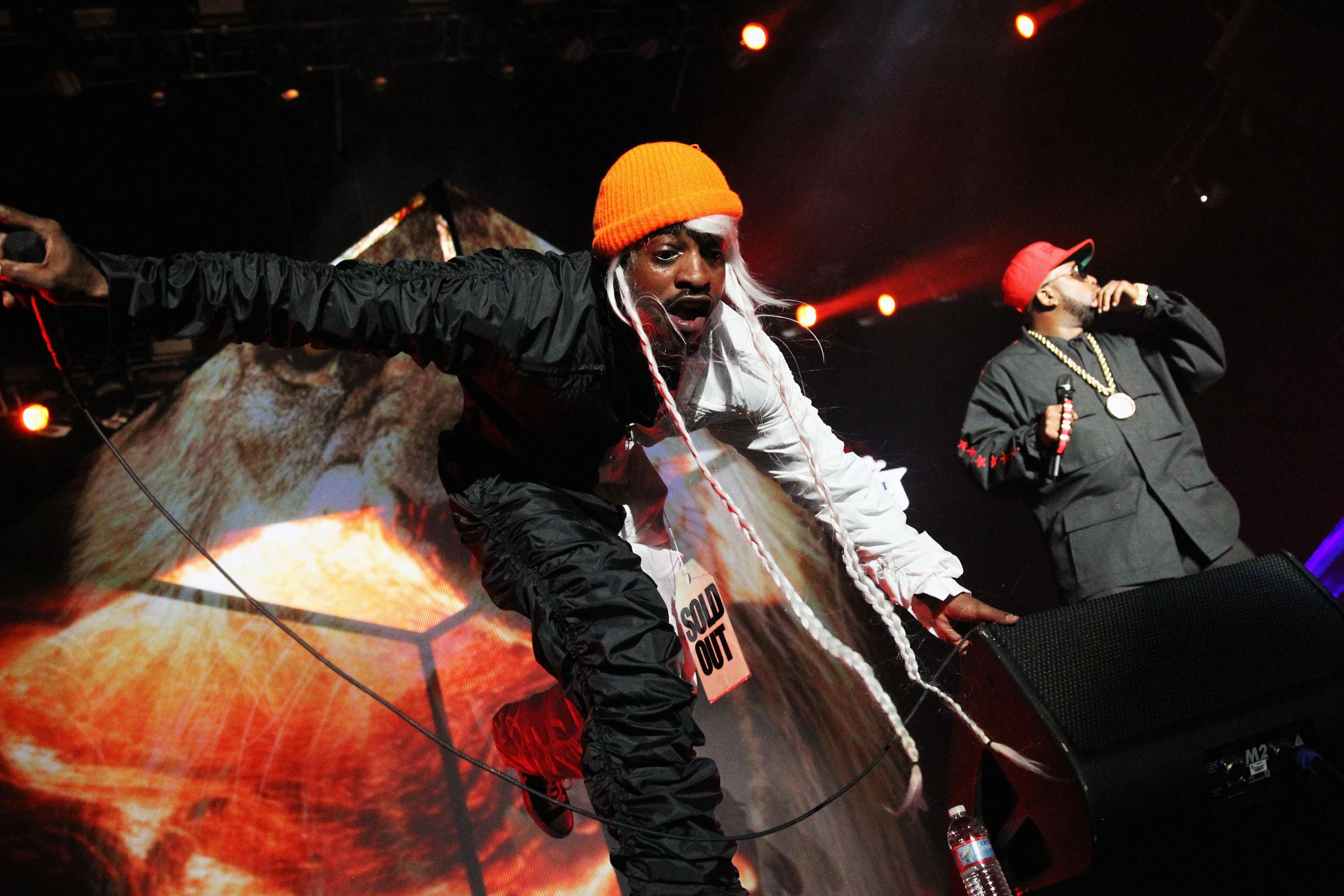 Two men in orange hats, black shirts, and black pants stand on a concert stage performing in front of red stage lights.