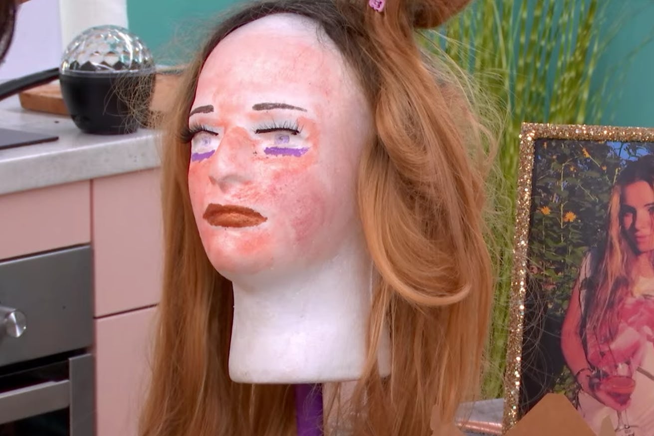 A mannequin head with a face of peach makeup and purple eyeliner wears a brown wig. Next to her is a framed photo of a girl with blonde hair holding flowers.