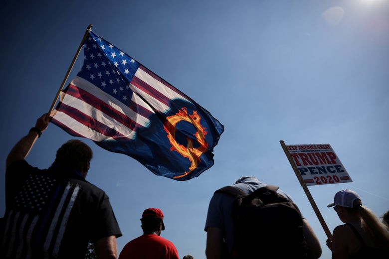 A man in a thin blue line t-shirt holds an American flag that is half emblazoned with a flaming Q beside another person holding a Trump-Pence 2020 sign, all seen from behind.