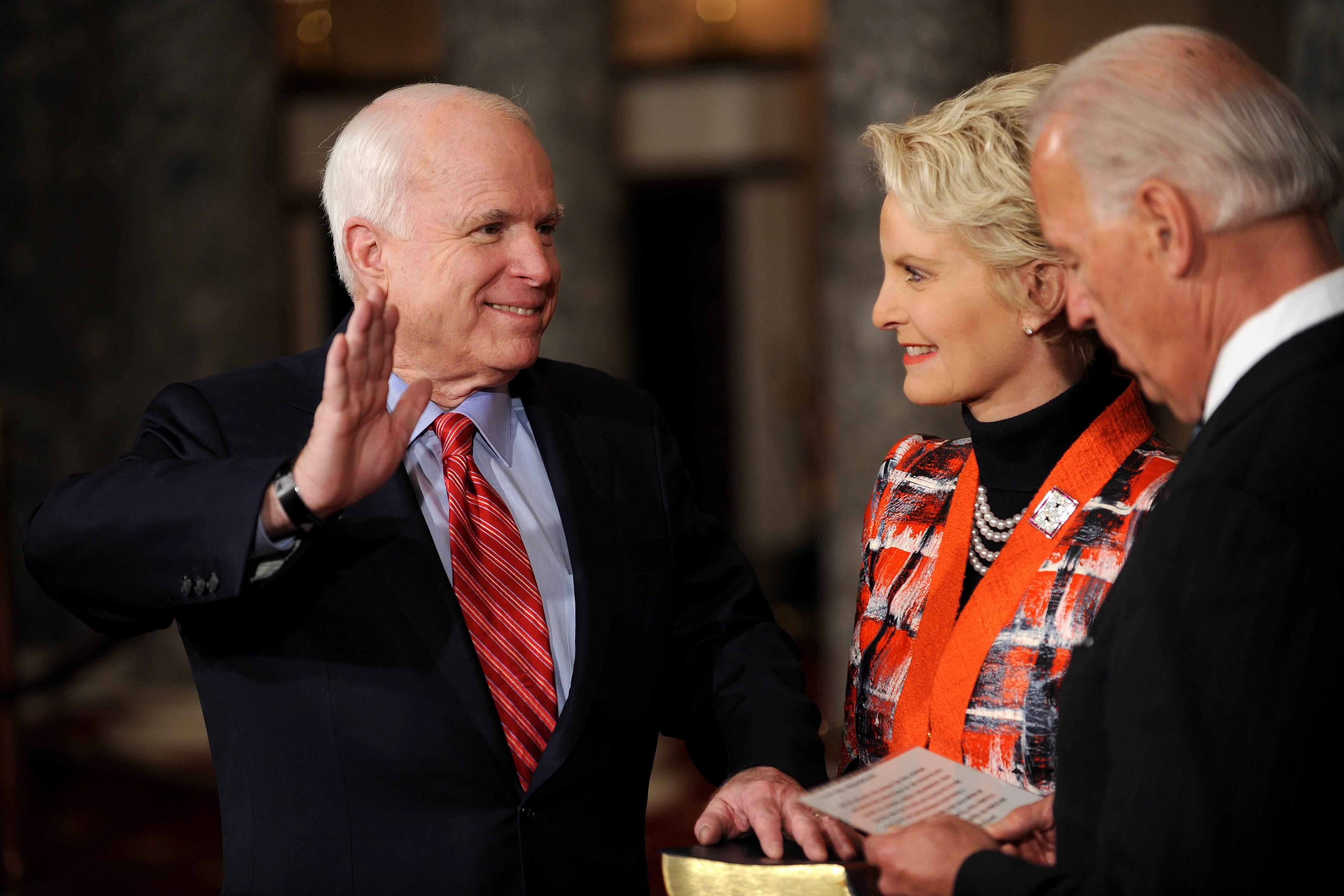 Cindy McCain holds a Bible on which John places his left hand as he raises his right hand. Joe Biden, in the foreground, reads from a piece of paper.