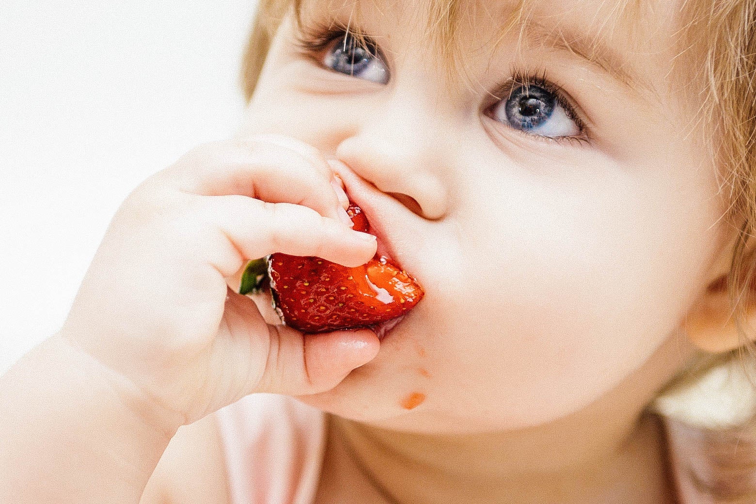 A toddler biting into a strawberry, making the juice dribble down their chin