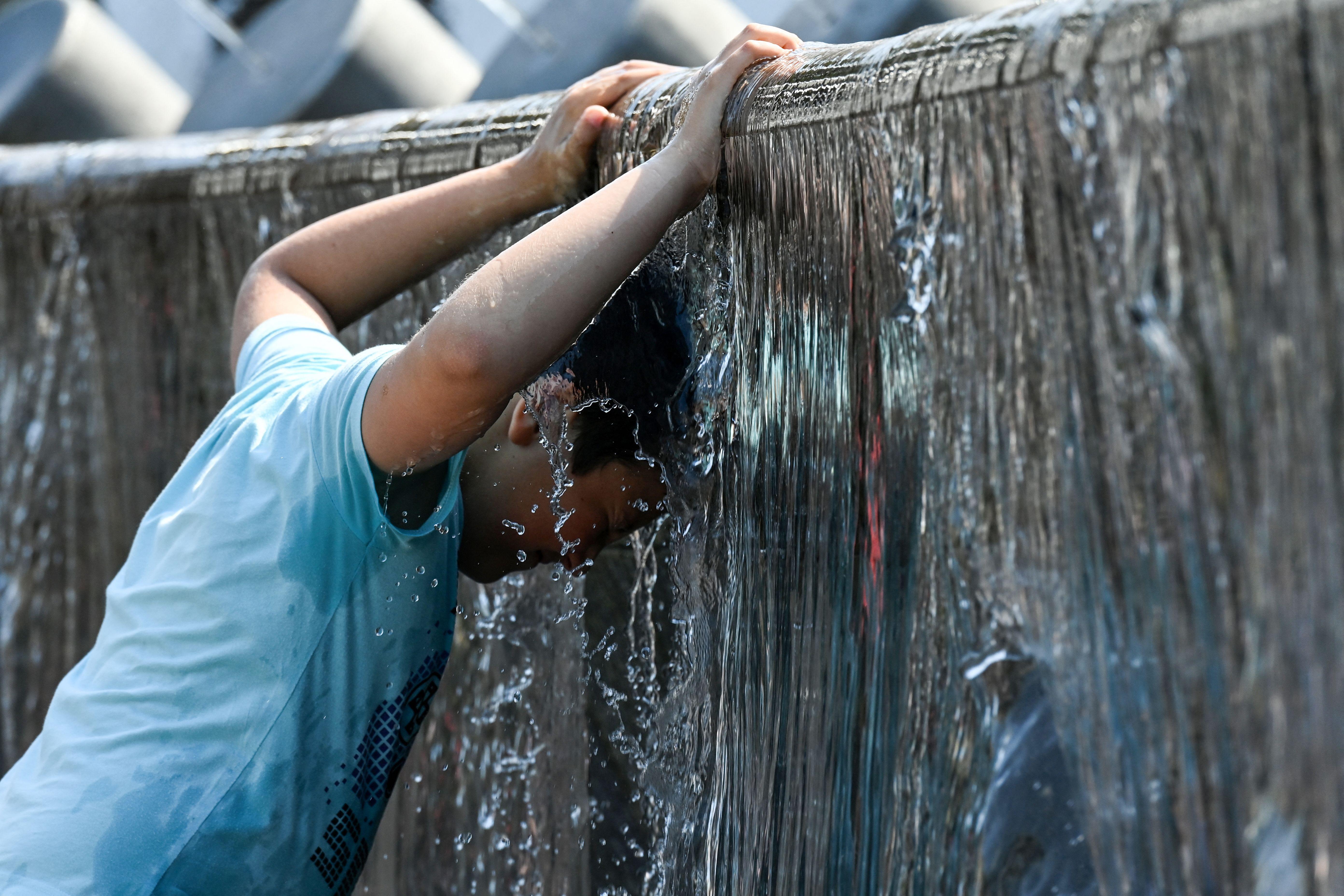 A boy in a blue T-shirt sticks his head under the water pouring off a fountain to cool off.