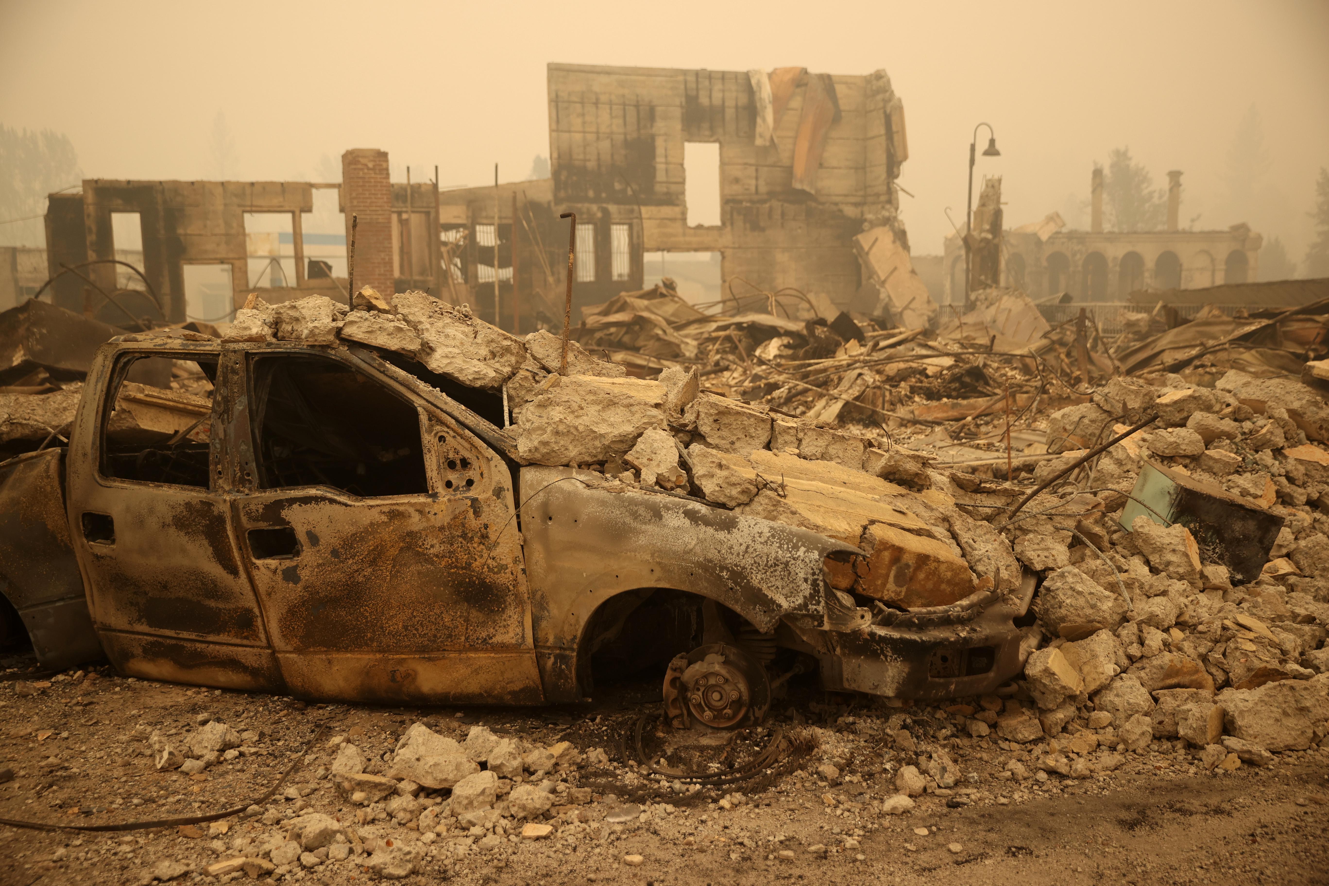 A burned truck is seen next to a pile of charred rubble and a damaged building.